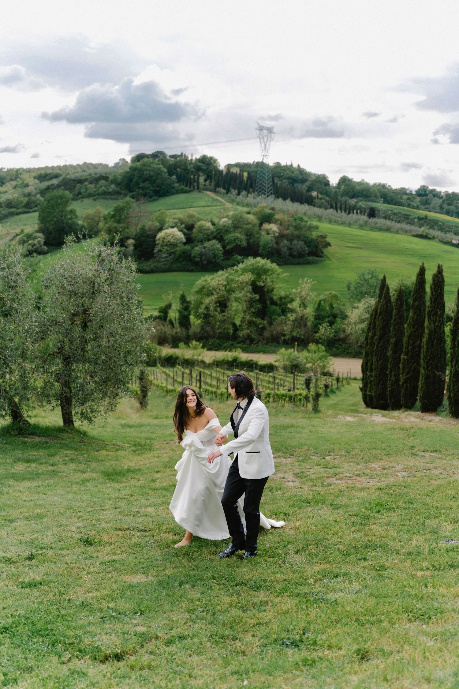 Bride and groom dance joyfully on green Tuscan hillside with rolling vineyards and cypress trees behind them.