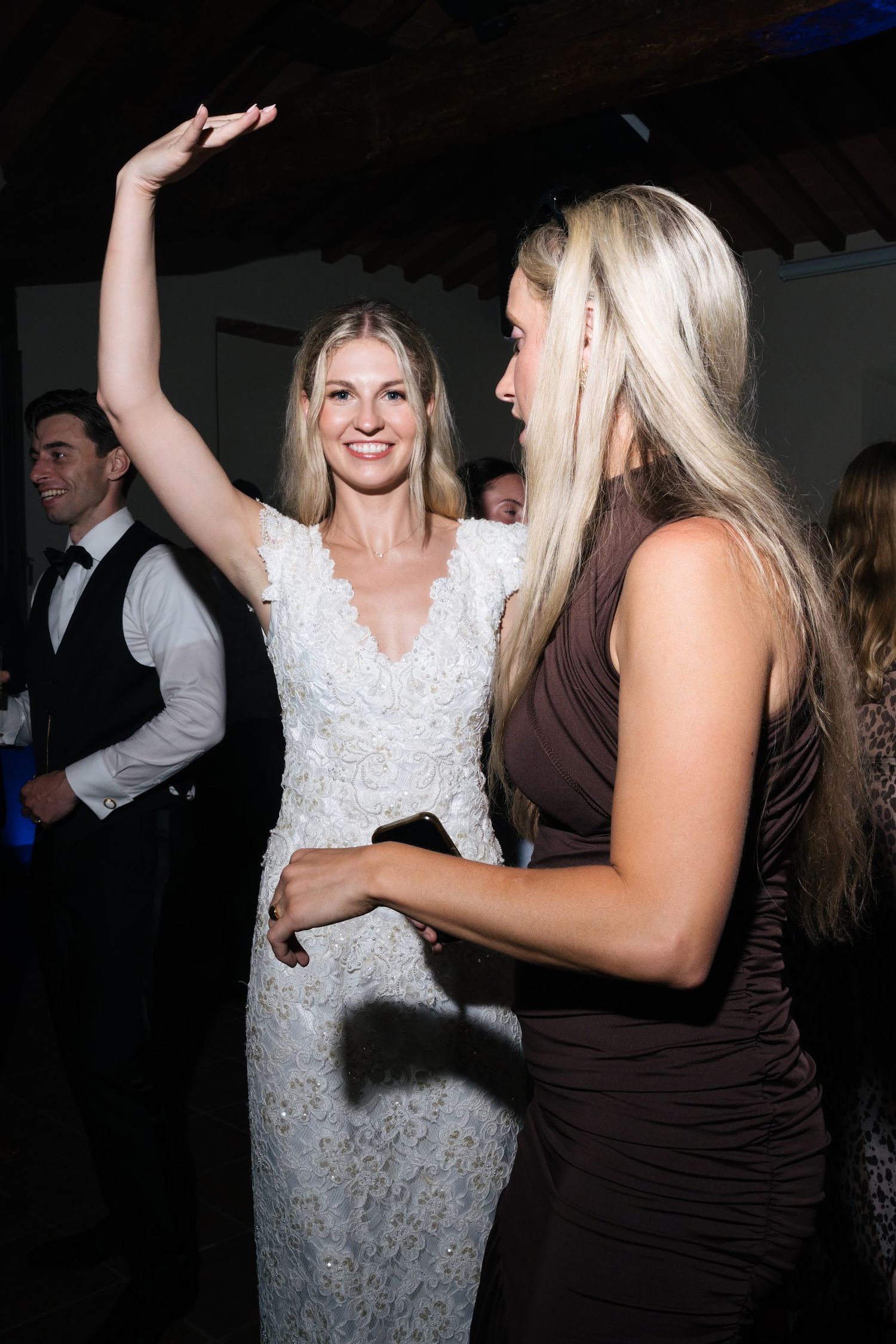 Smiling bride in white lace gown raises arm while dancing with guest at reception.