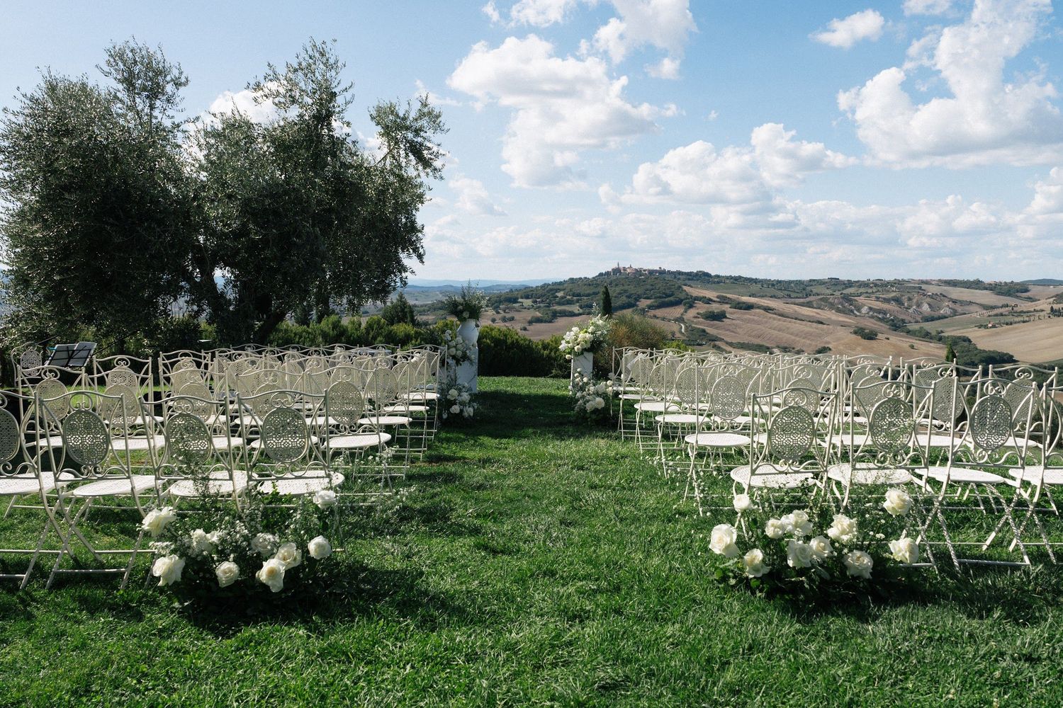 Elegant outdoor wedding ceremony setup with white chairs and floral arrangements on green lawn with Tuscan hills backdrop.