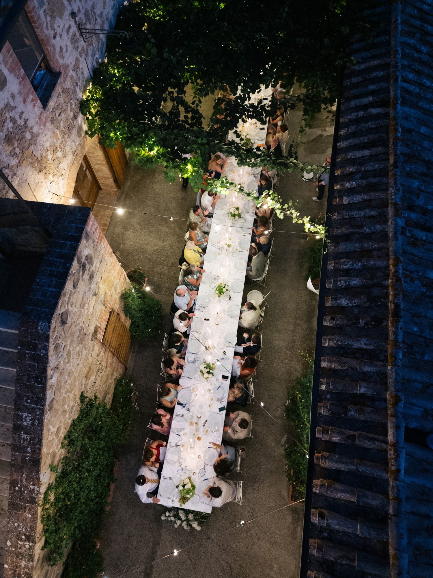 Aerial night view of wedding guests dining at long illuminated table in rustic stone courtyard.
