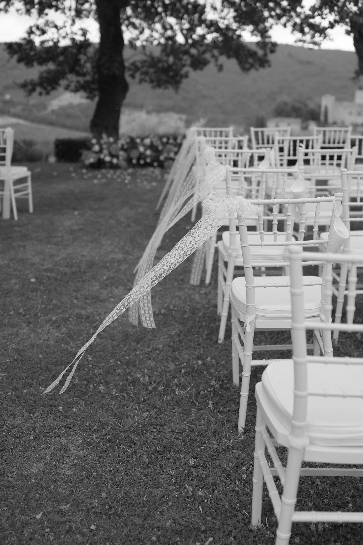 Black and white photo of white Chiavari chairs with lace ribbon decorations at an outdoor wedding ceremony.
