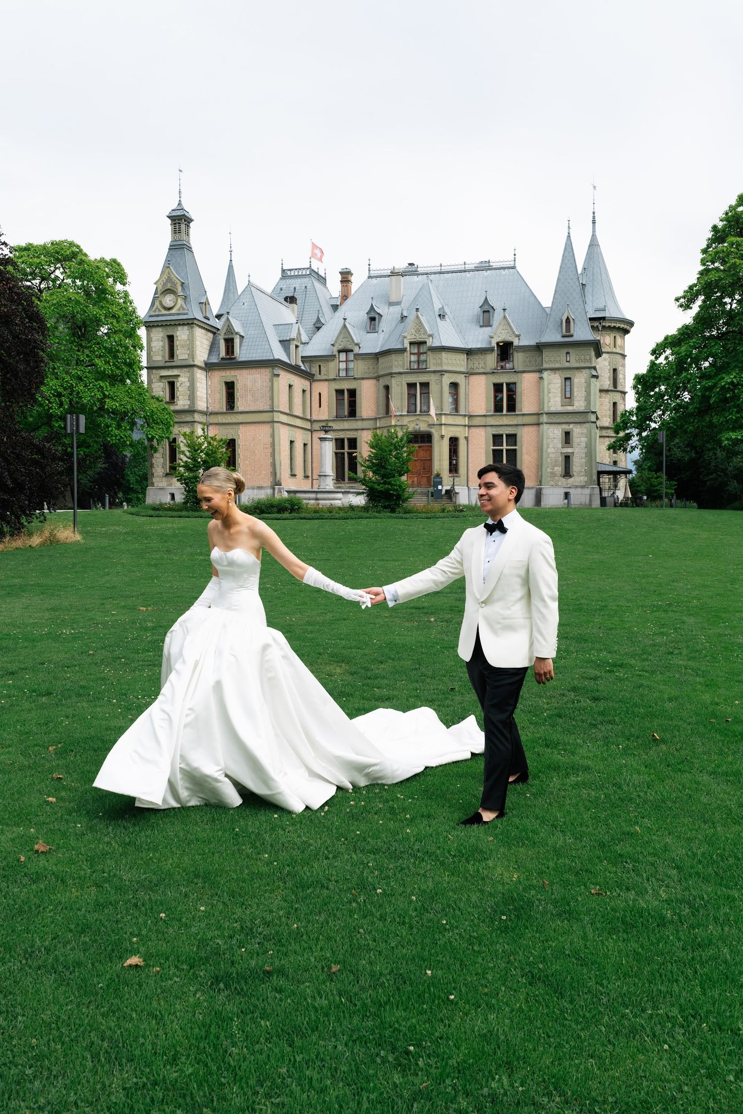 Bride and groom holding hands walking across green lawn in front of a grand château wedding venue.