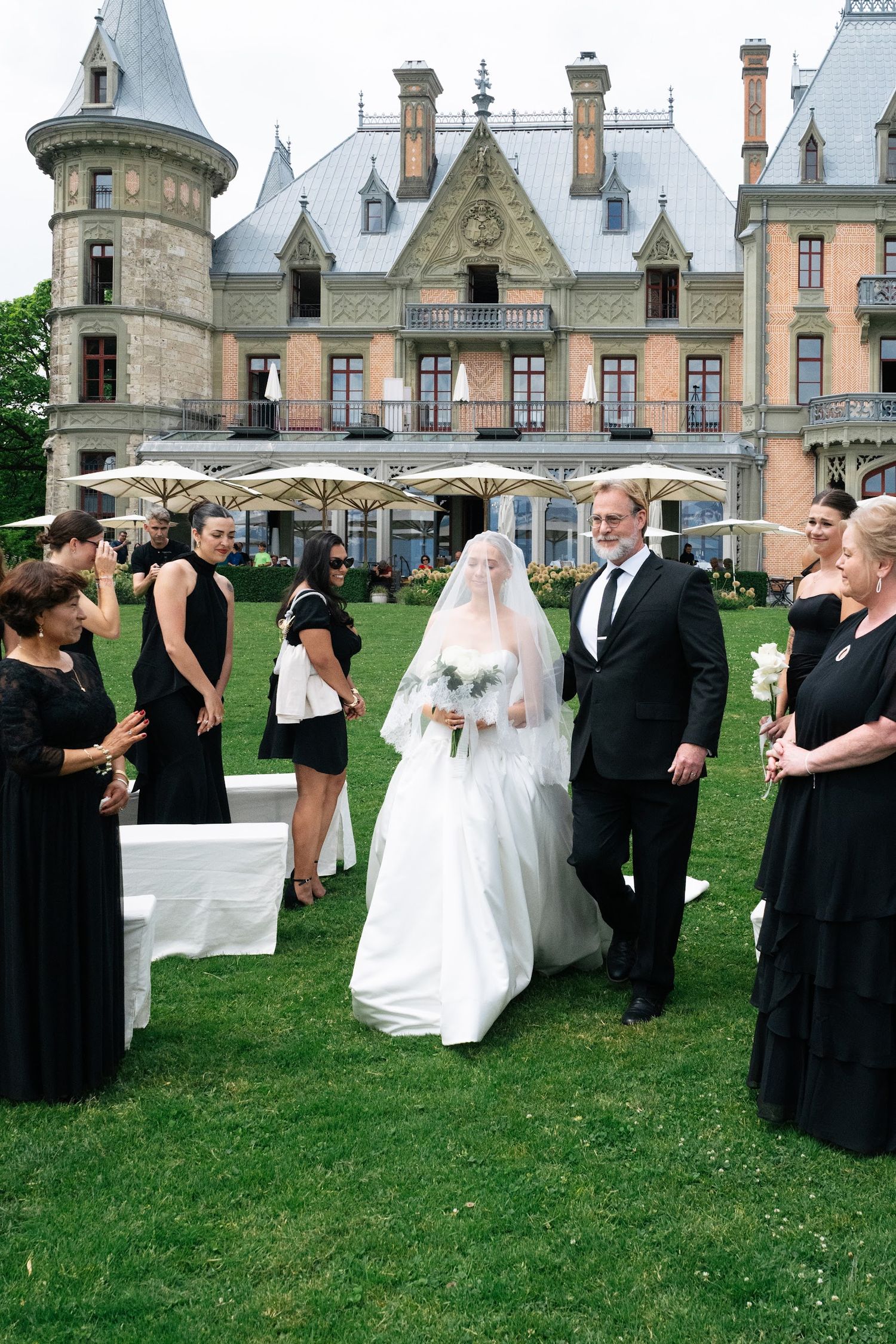 Bride in a white gown and long veil walks down the aisle escorted by her father at an outdoor château wedding.