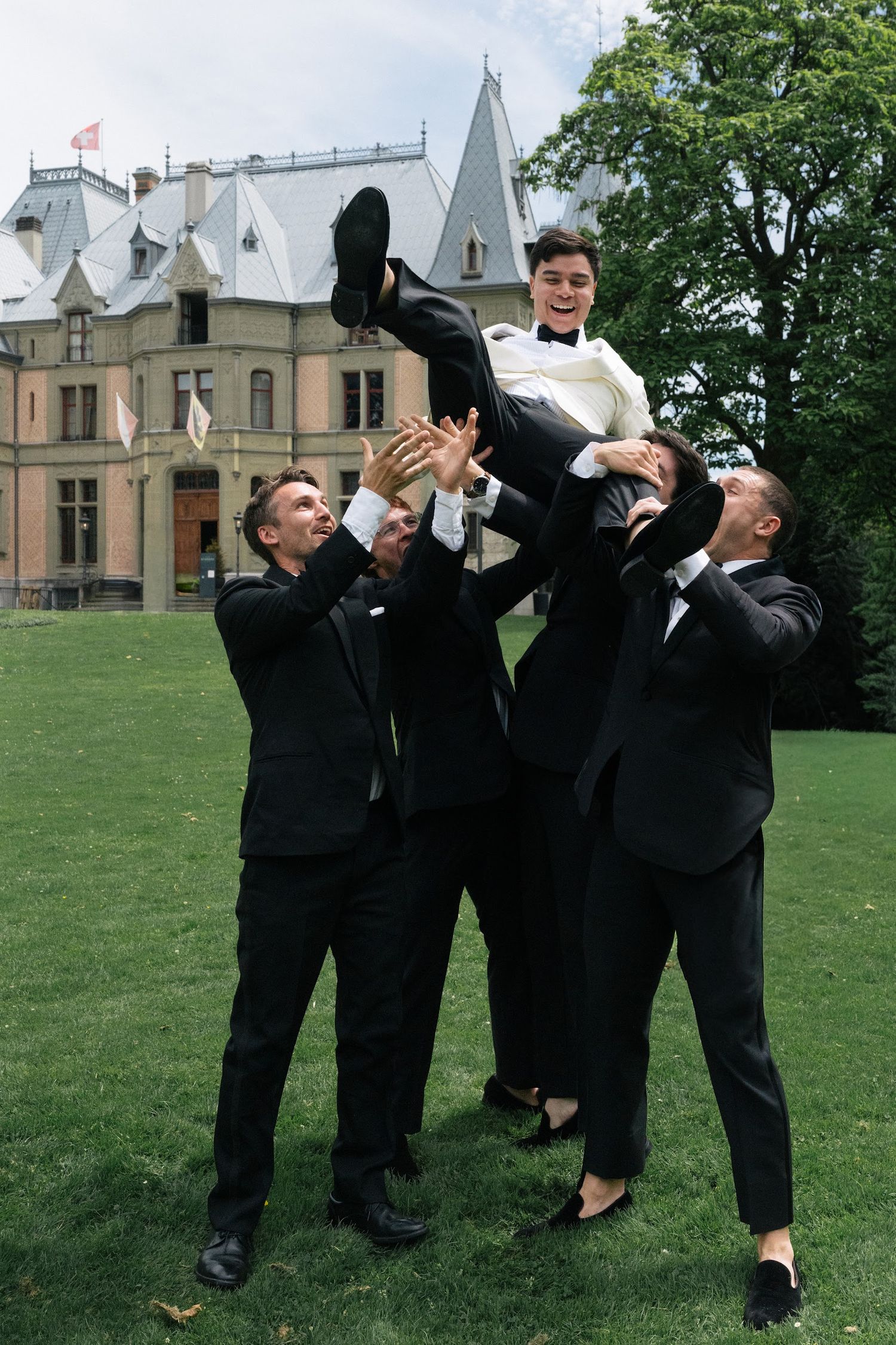 Groomsmen lifting laughing groom in white tuxedo into air on green lawn outside castle venue.
