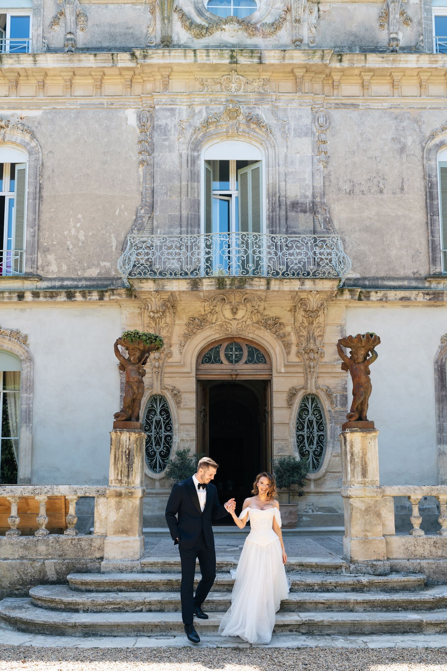 Bride and groom hold hands on steps of an ornate baroque chateau with classical stone sculptures.