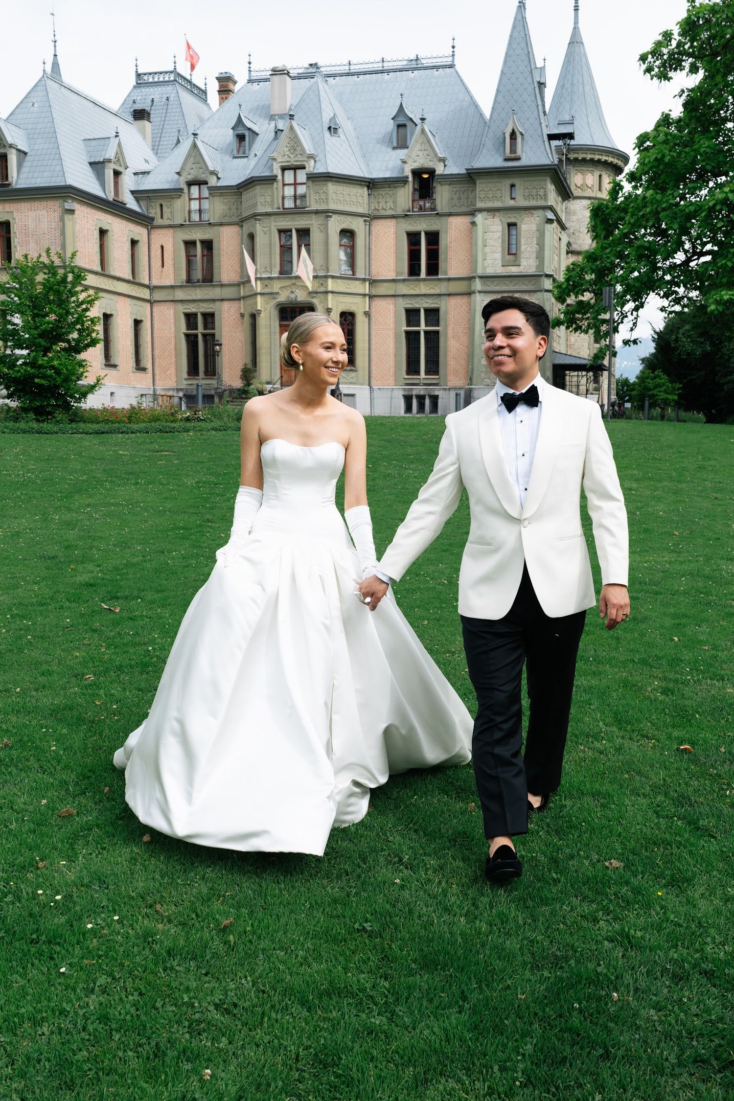 Bride and groom walking hand in hand smiling before a grand castle venue with Swiss flag flying.