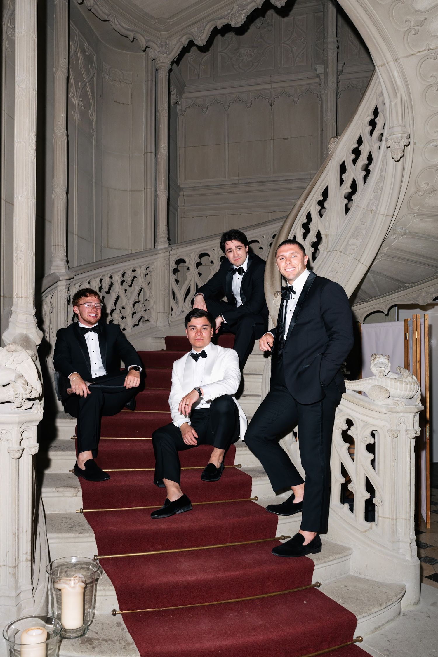 Four groomsmen in black tuxedos posing on ornate white marble staircase with red carpet runner.