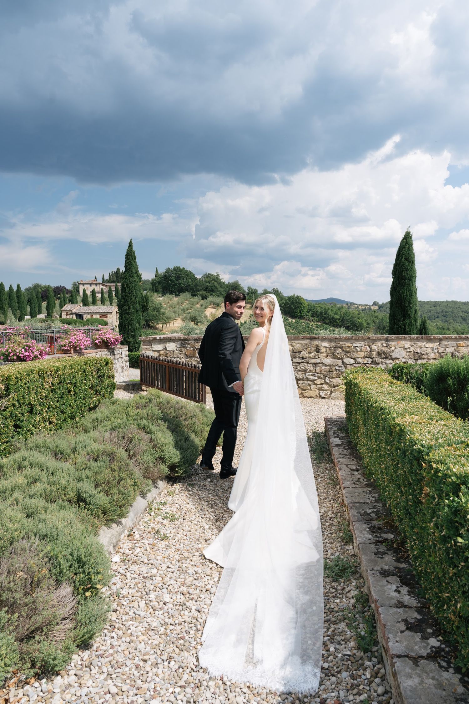 Bride in a long white gown with cathedral veil stands with groom in formal gardens under dramatic skies.