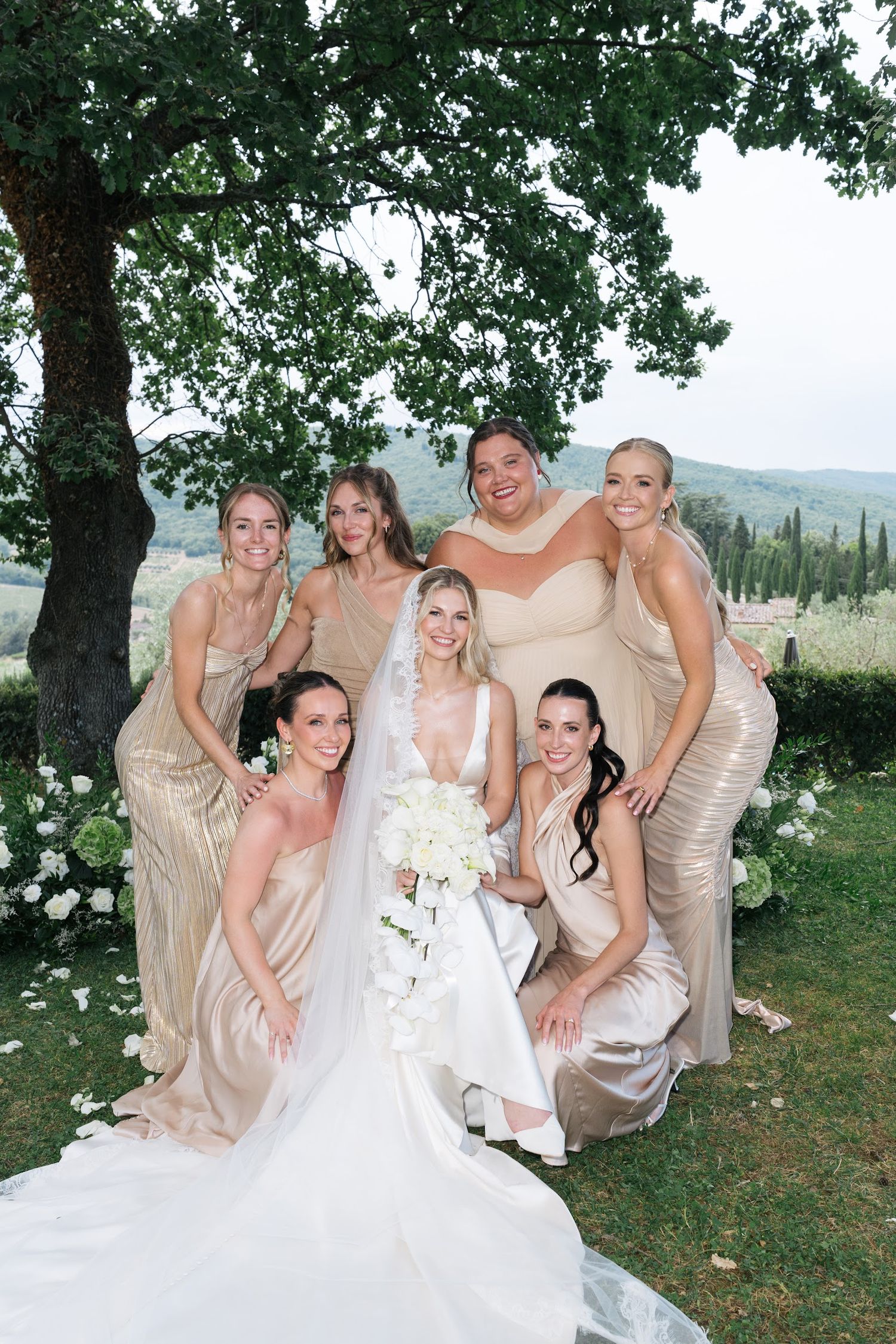 Bride poses with six bridesmaids in champagne gowns under a tree with Tuscan hills behind them.