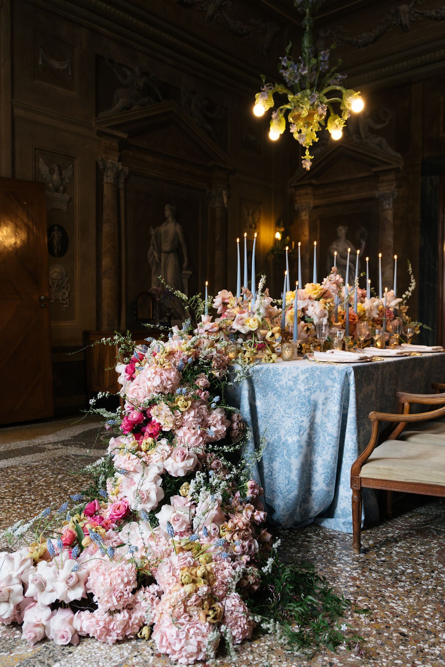 Lavish wedding reception table adorned with cascading pink peonies, tall candles, and blue linen in ornate villa.
