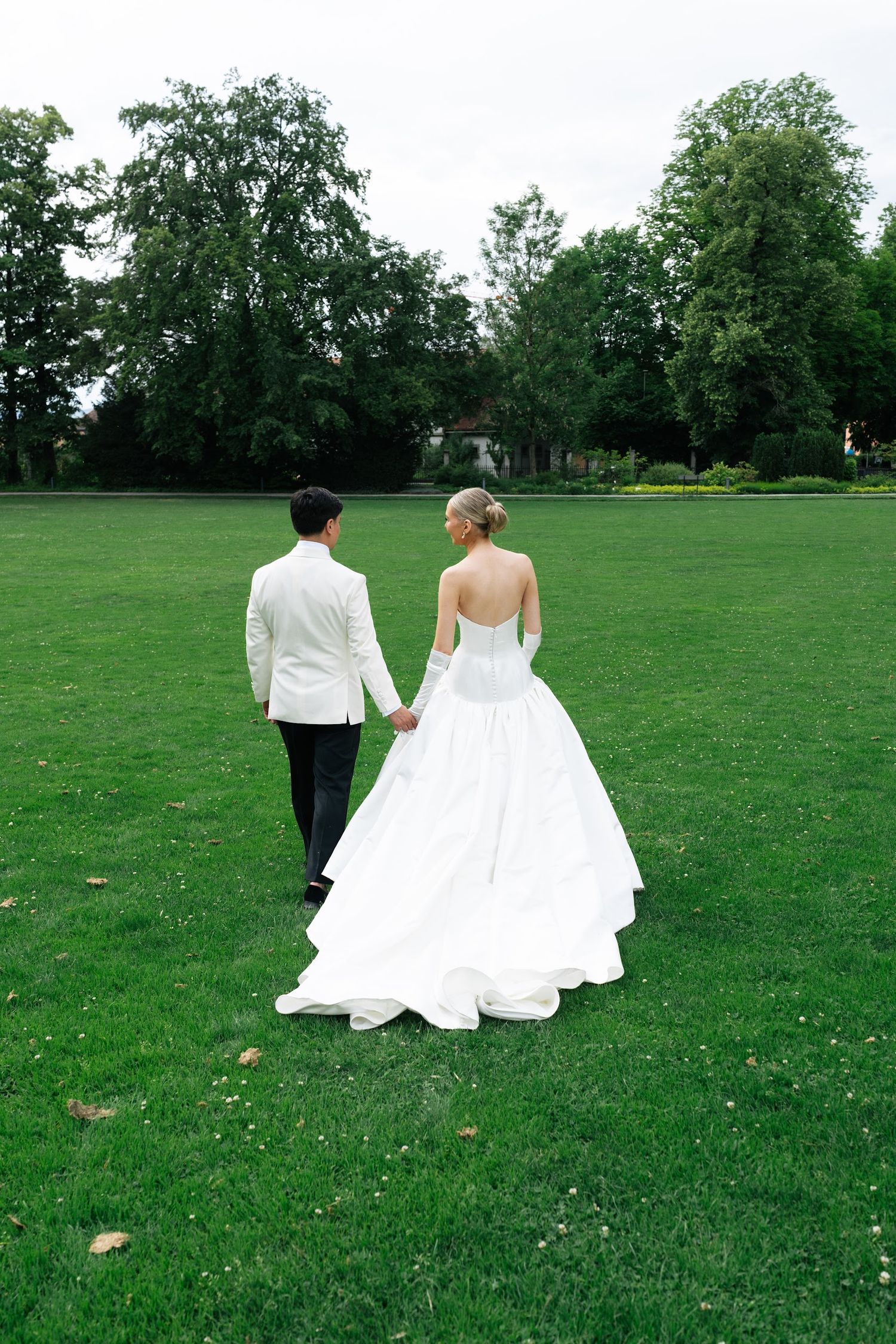 Bride and groom walking hand in hand away from camera across expansive green lawn with trees.
