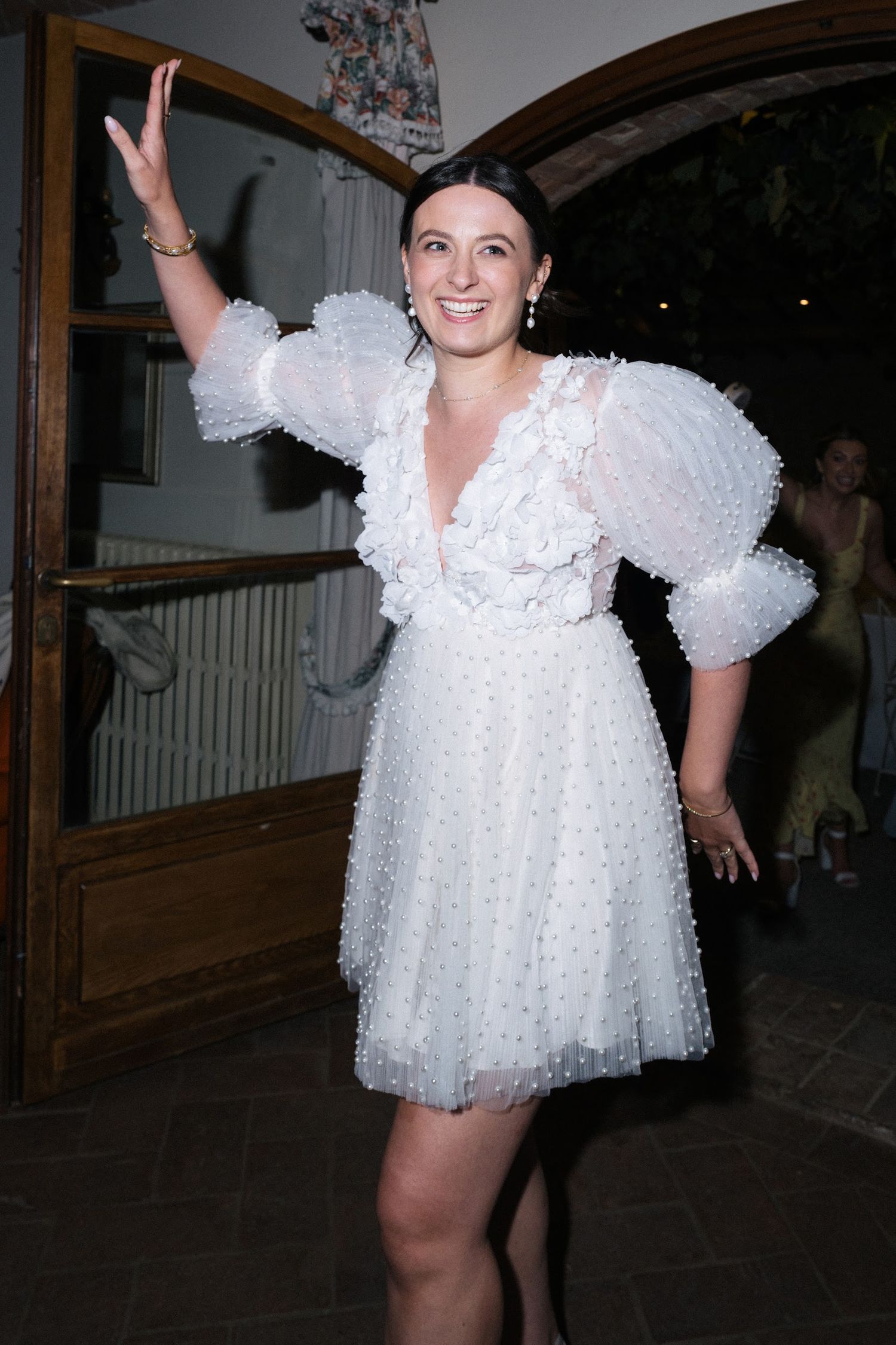 Bride in white ruffled pearl mini dress waving at wedding reception.
