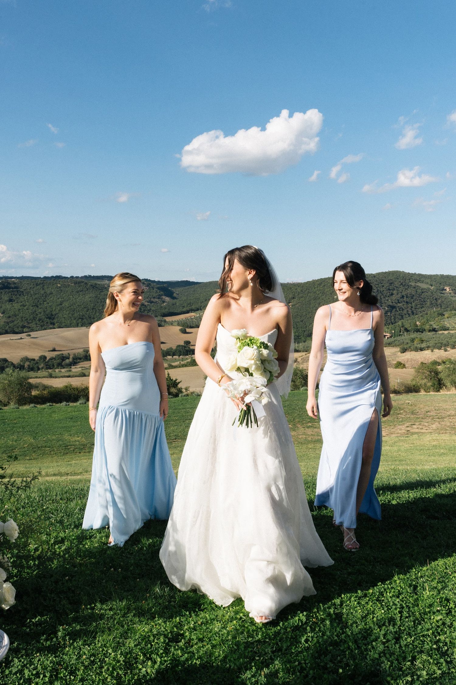 Bride in white gown walks with two bridesmaids in light blue dresses across green Tuscan hillside.