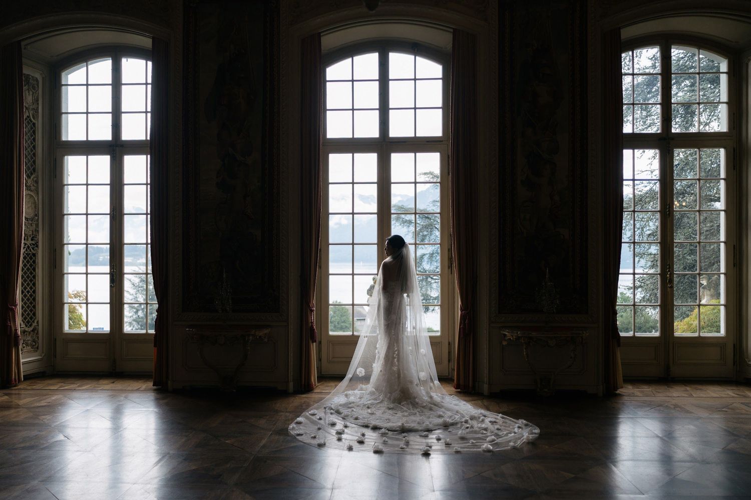 Bride in cathedral veil stands before grand arched windows in ornate ballroom with mountain lake views.