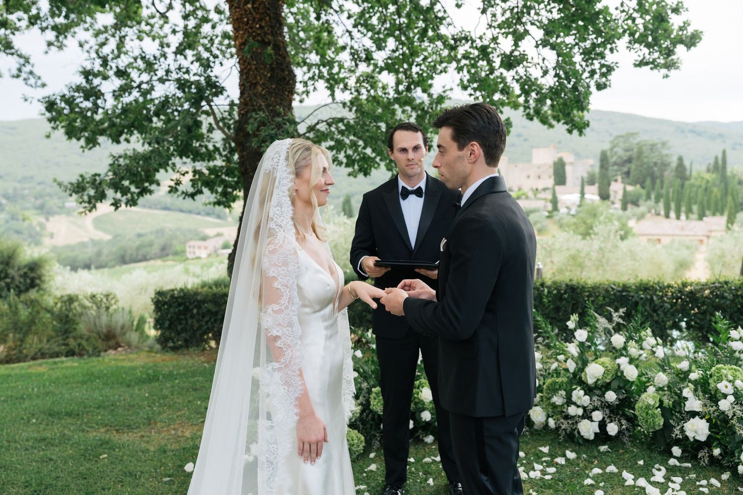 Groom places ring on bride's finger during outdoor wedding ceremony in Tuscany with cypress trees.