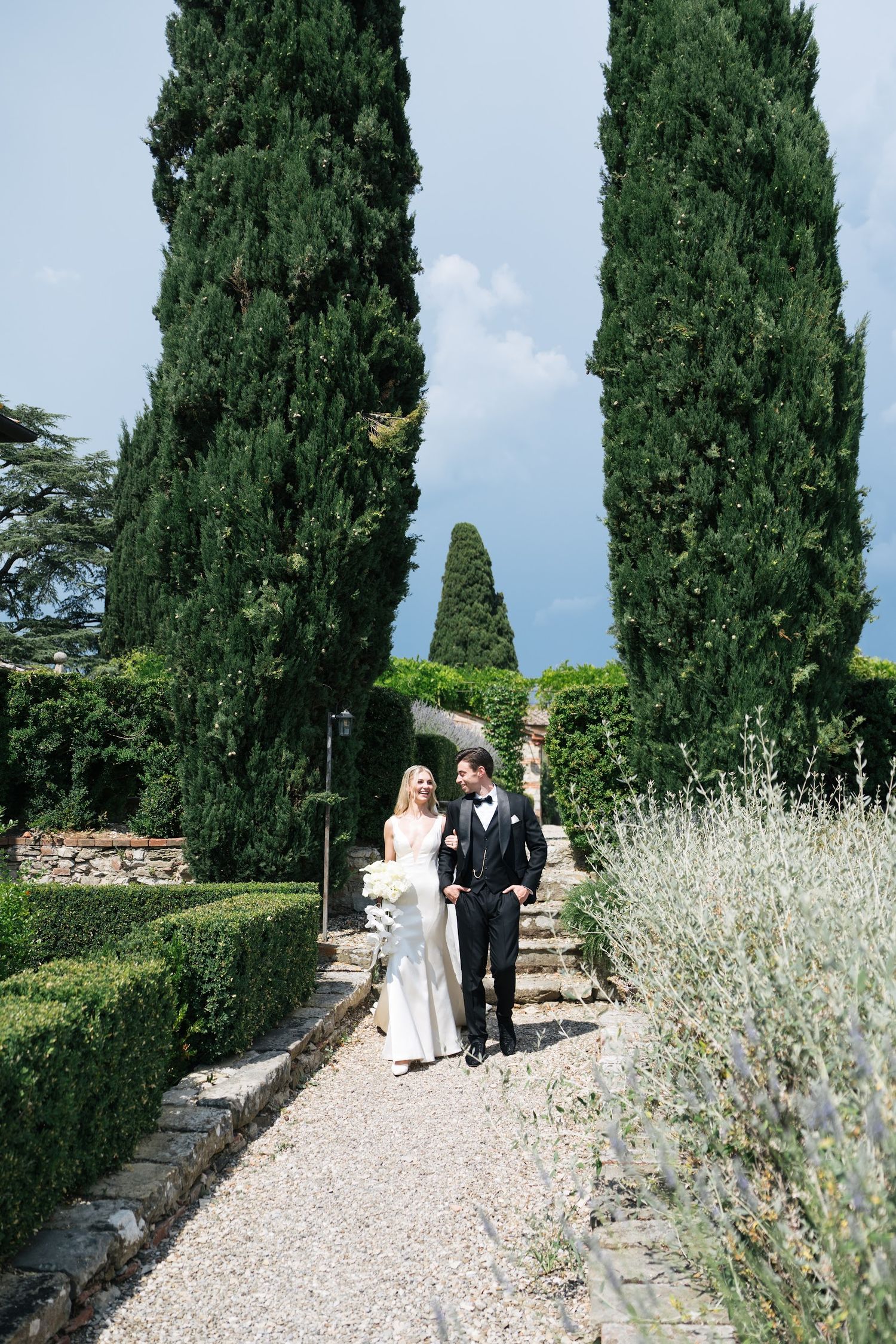 Bride and groom walk hand in hand along a gravel path lined with cypress trees and boxwood hedges.