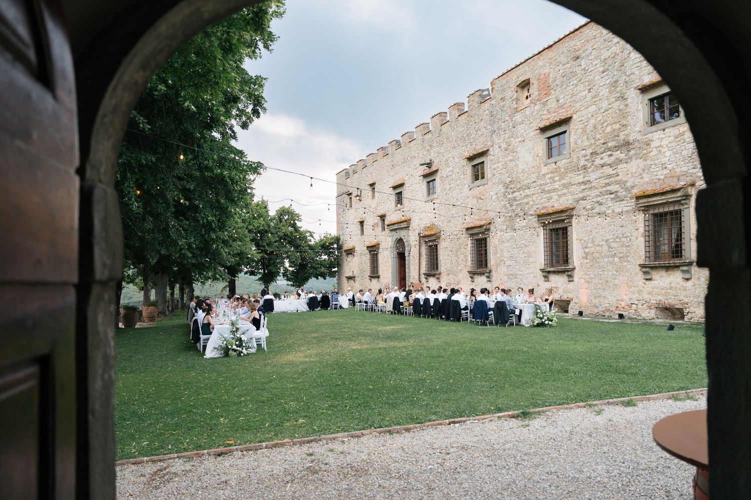 Outdoor wedding reception on green lawn viewed through stone archway of historic villa.