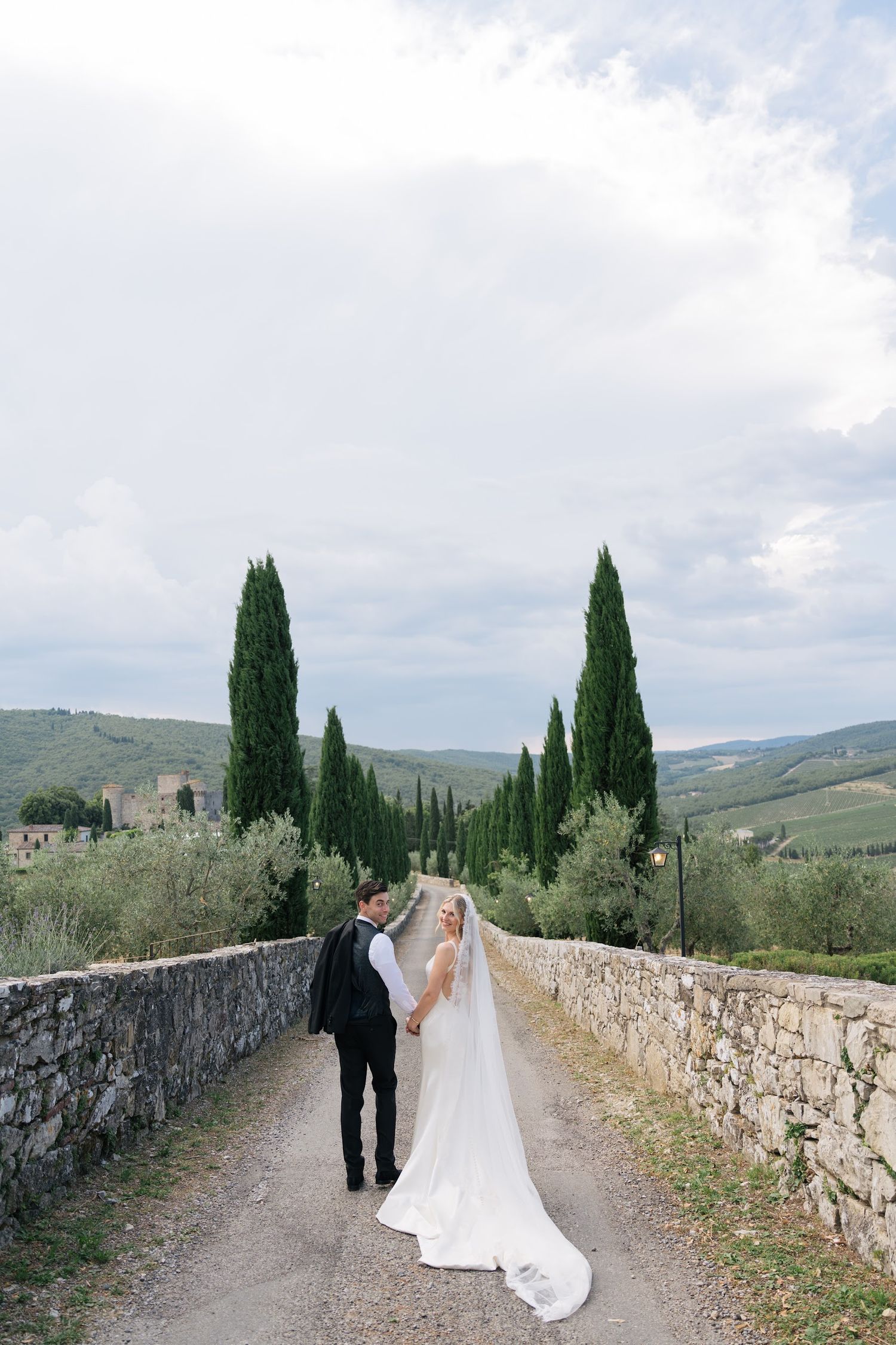 Bride and groom walk hand-in-hand on a stone path lined with cypress trees in the Tuscan countryside.