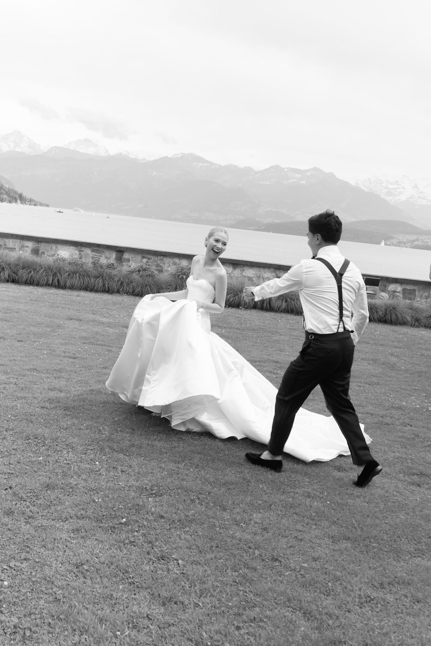 Black and white photo of bride and groom dancing on lawn with mountain lake scenery behind them.