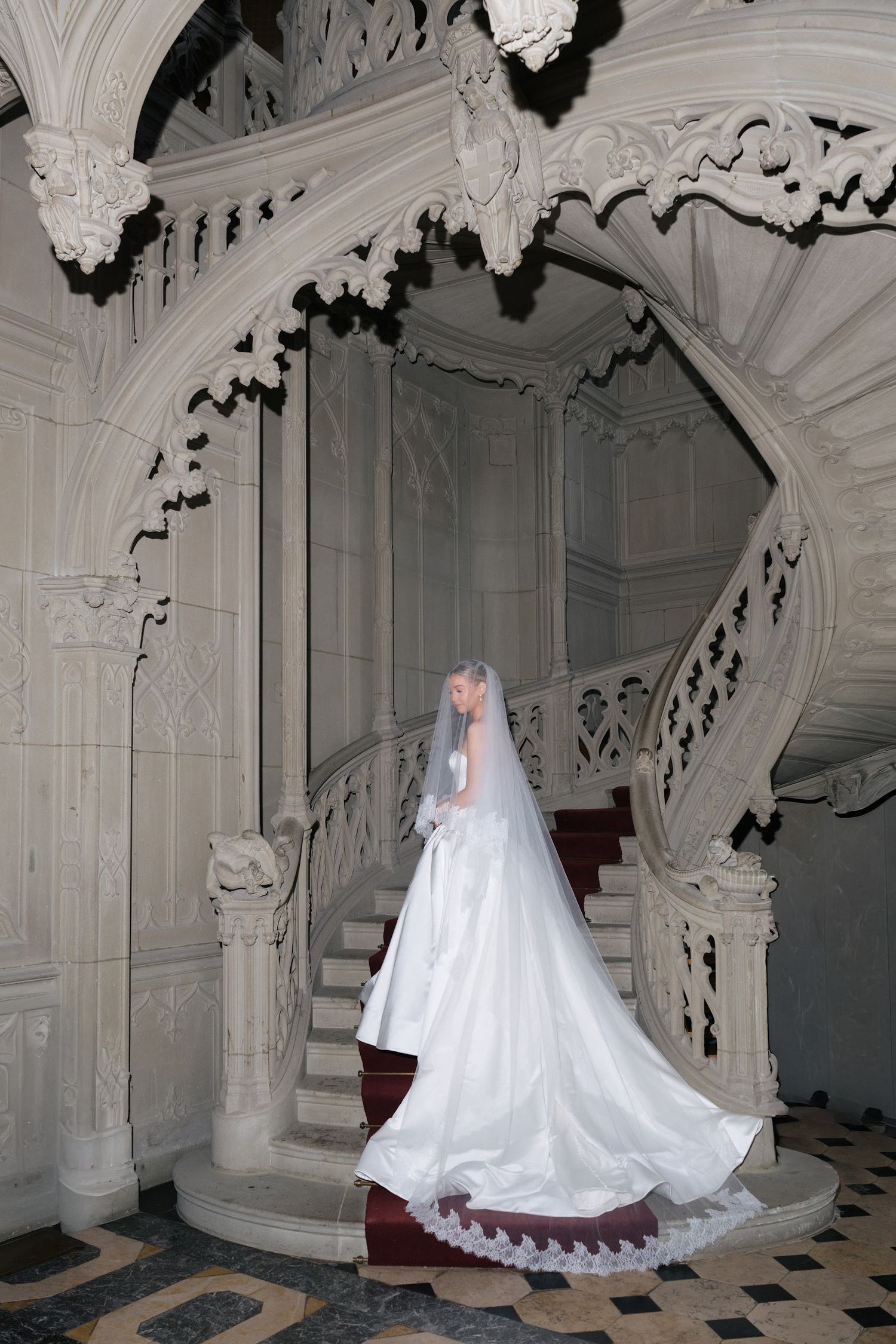 Elegant bride in a long veil and satin gown poses on an intricate white Gothic spiral staircase.