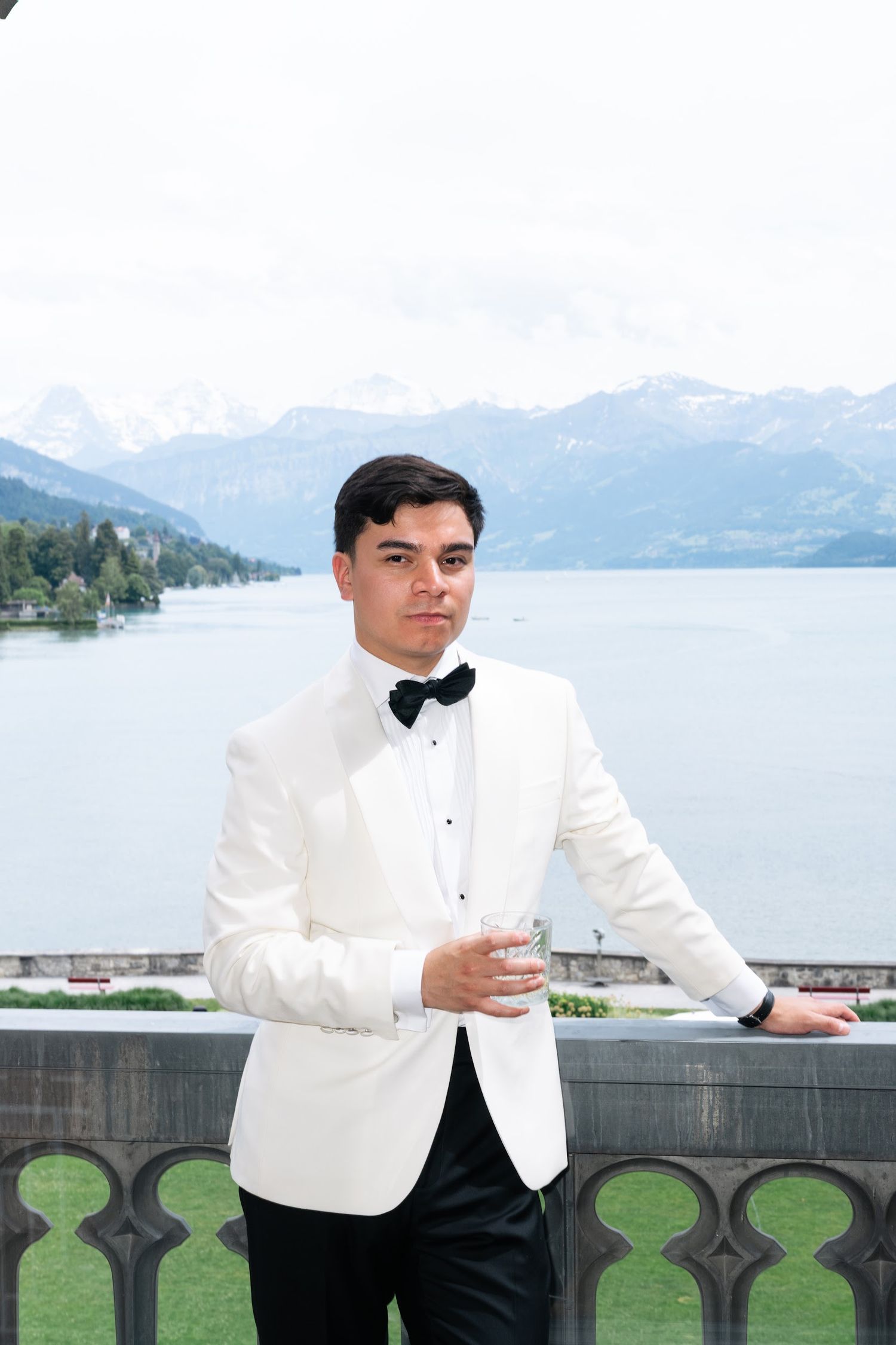 Groom in white dinner jacket and black bow tie poses on balcony overlooking scenic alpine lake and mountain landscape.
