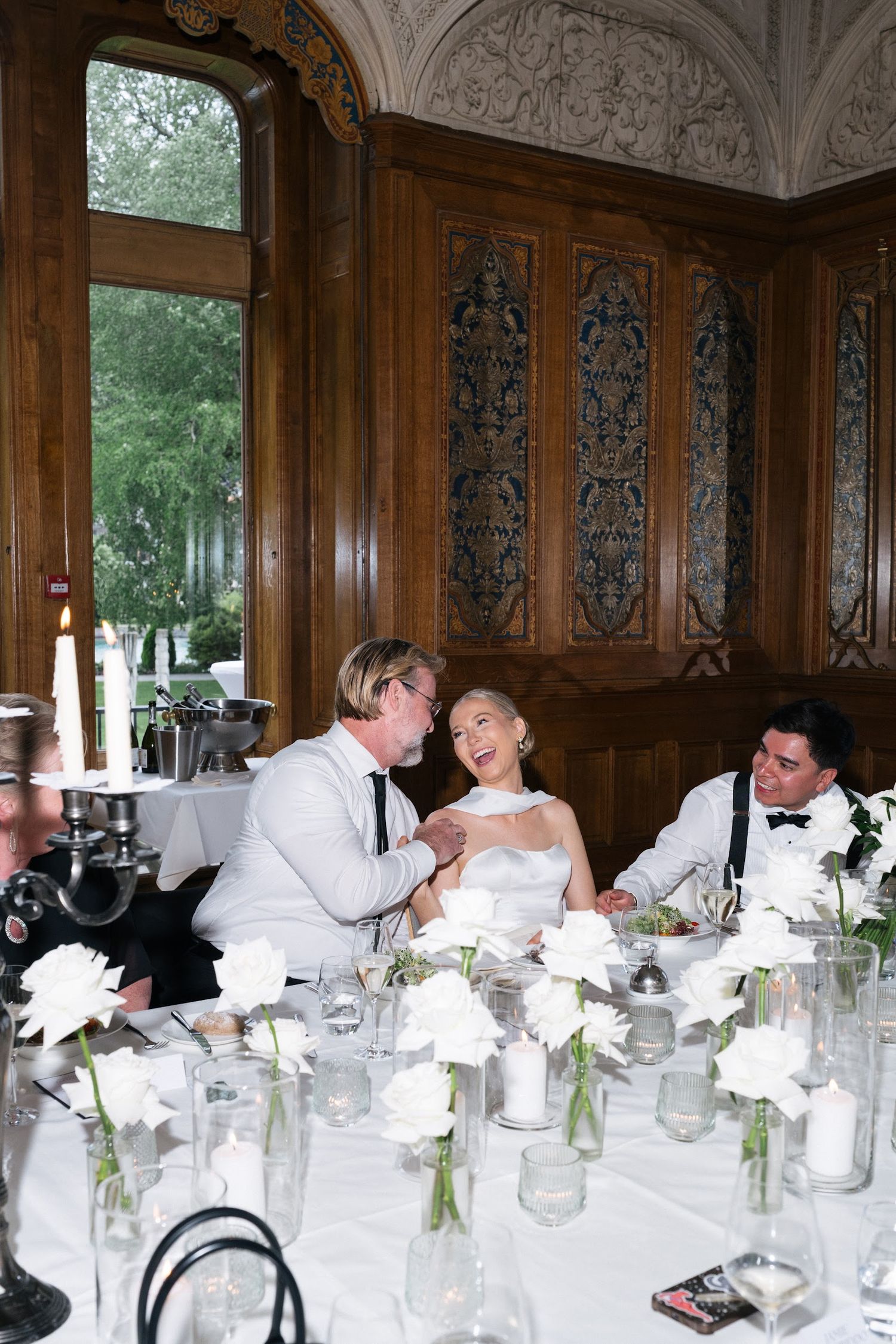 Bride in white gown laughs with guests at ornate wood-paneled wedding reception venue.