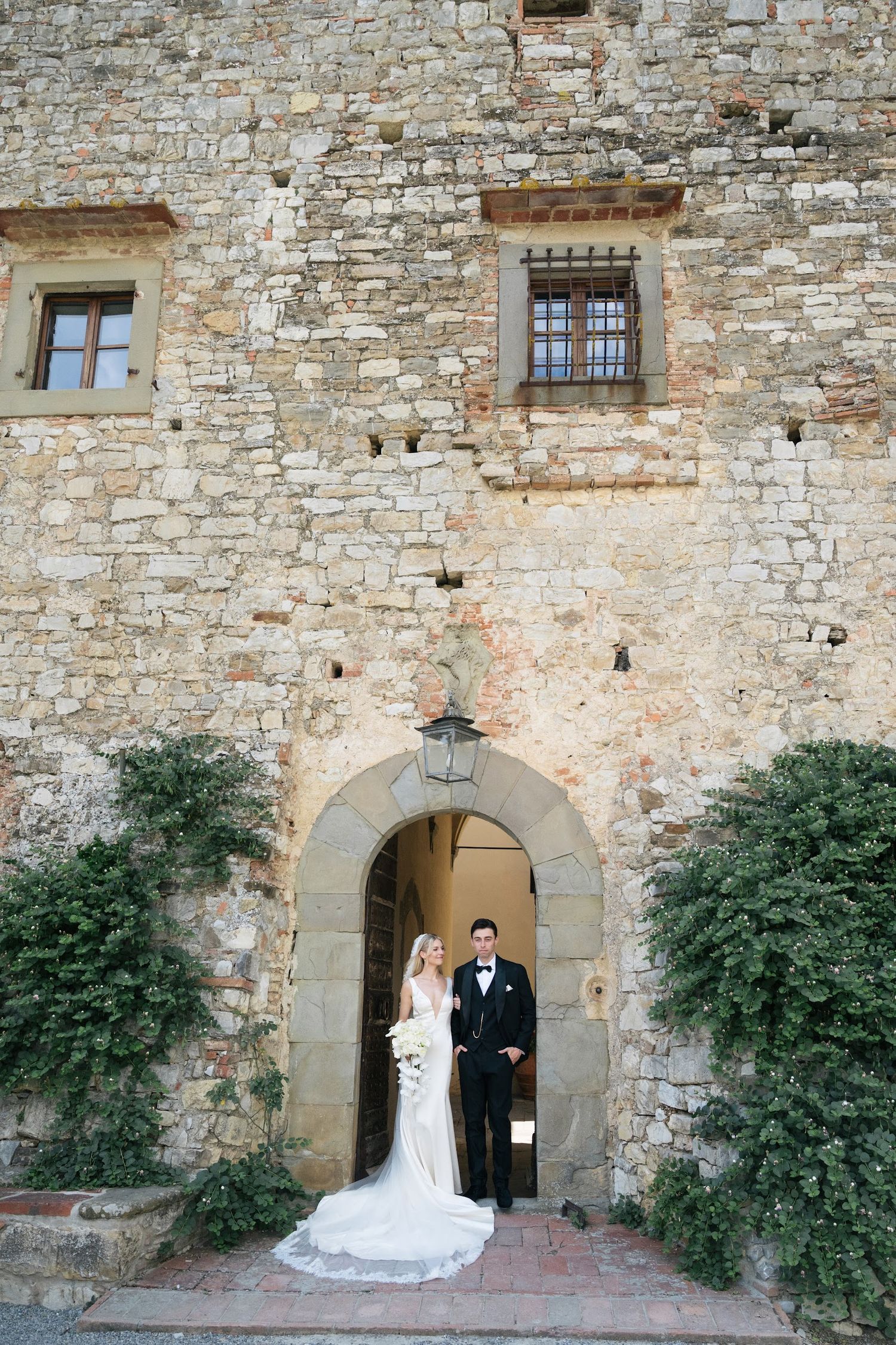 A bride and groom pose beneath a stone archway of a rustic Tuscan castle wedding venue.