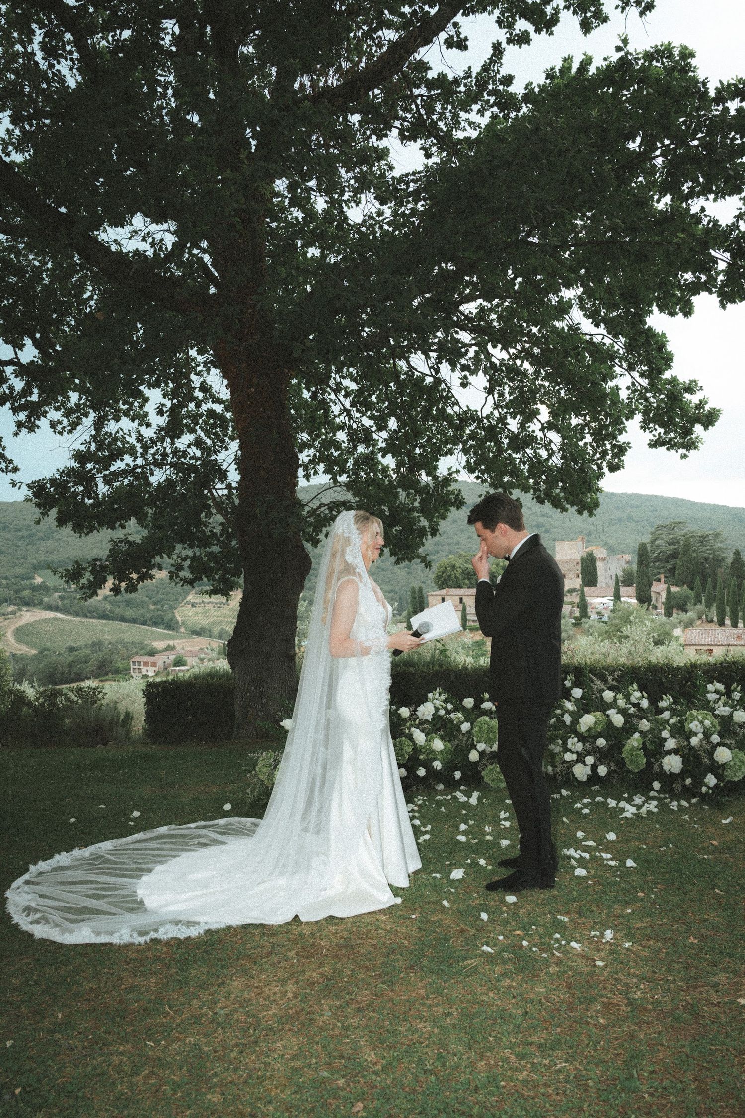 Groom in black tuxedo reads vows to bride in white gown under oak tree with Tuscan landscape backdrop.