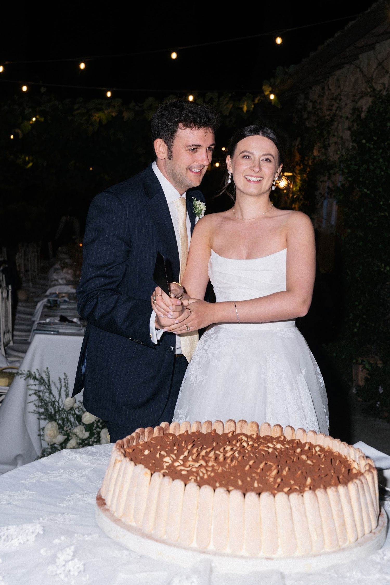 Bride and groom smile while cutting their tiramisu wedding cake outdoors.
