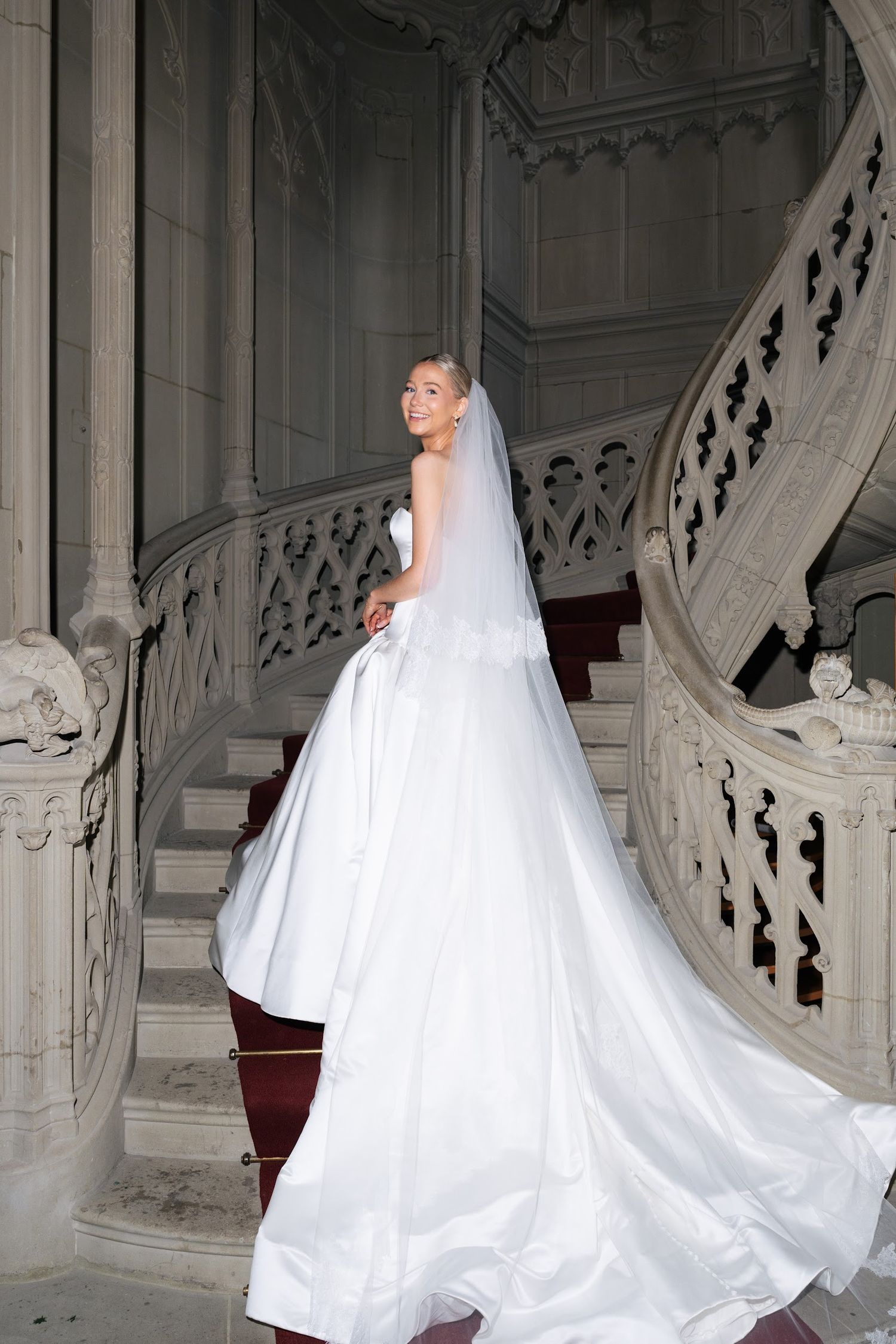 Elegant bride in white ball gown and veil posing on grand ornate marble staircase.