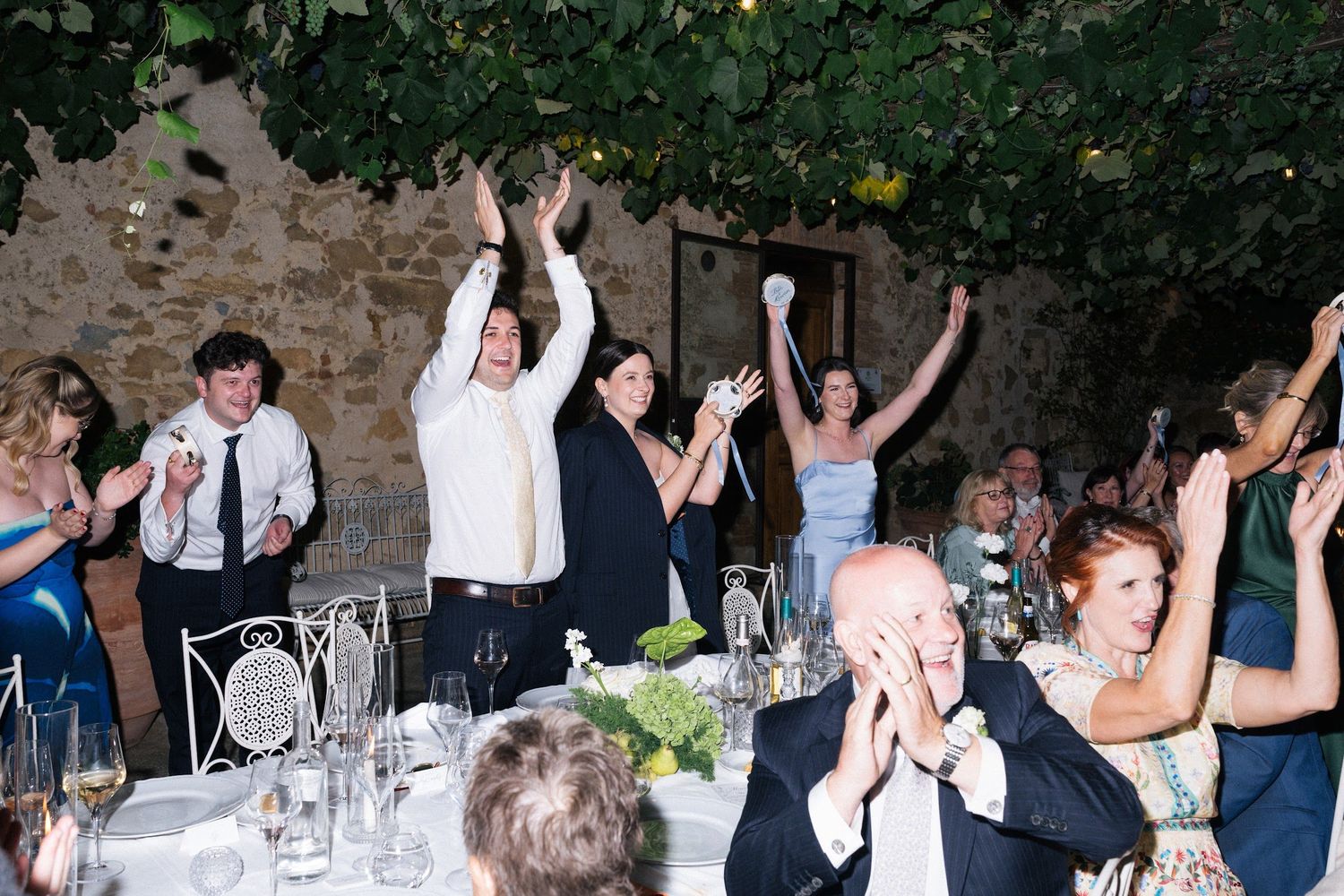 Wedding guests cheer with hands raised at an elegant outdoor reception dinner.