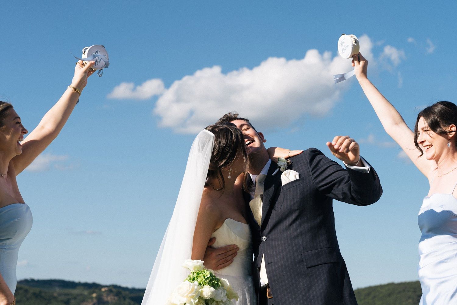 Joyful bride and groom kiss as bridesmaids in blue dresses raise white doves at outdoor Tuscany wedding.