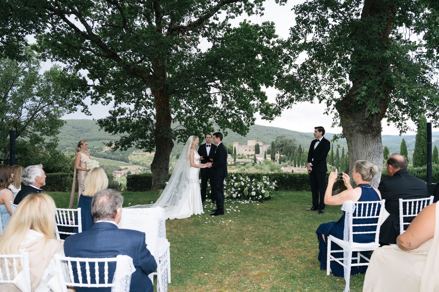 Bride in white gown and veil meets groom at altar under oak tree during Tuscan outdoor ceremony.
