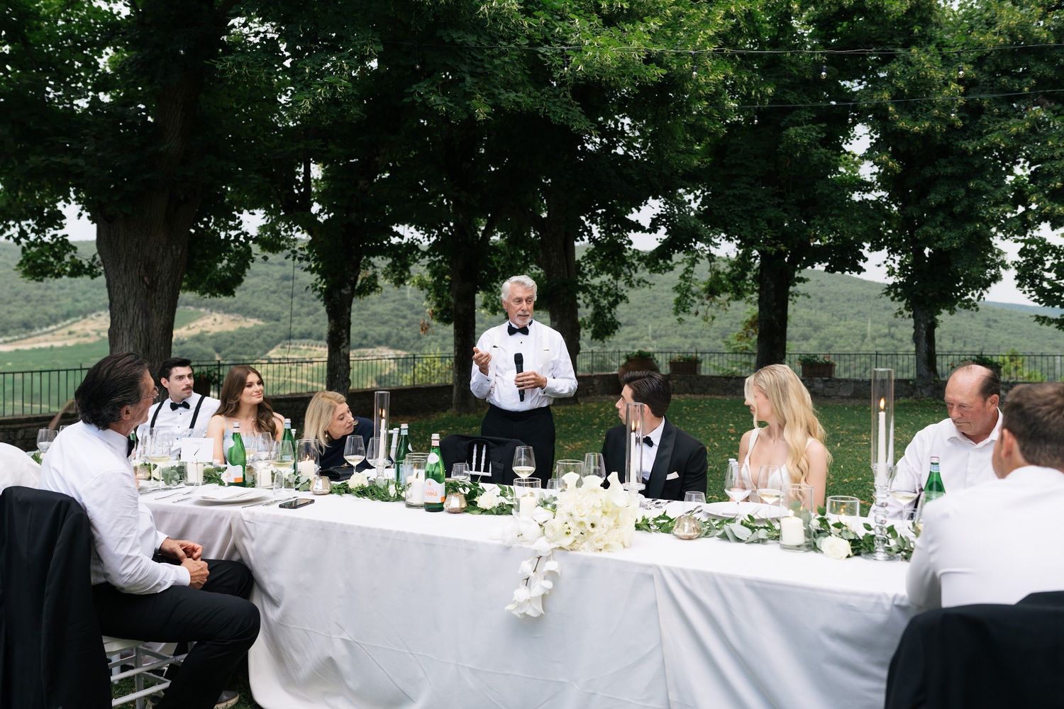Wedding speaker delivers a toast at an elegant outdoor Tuscan reception with rolling hills backdrop.