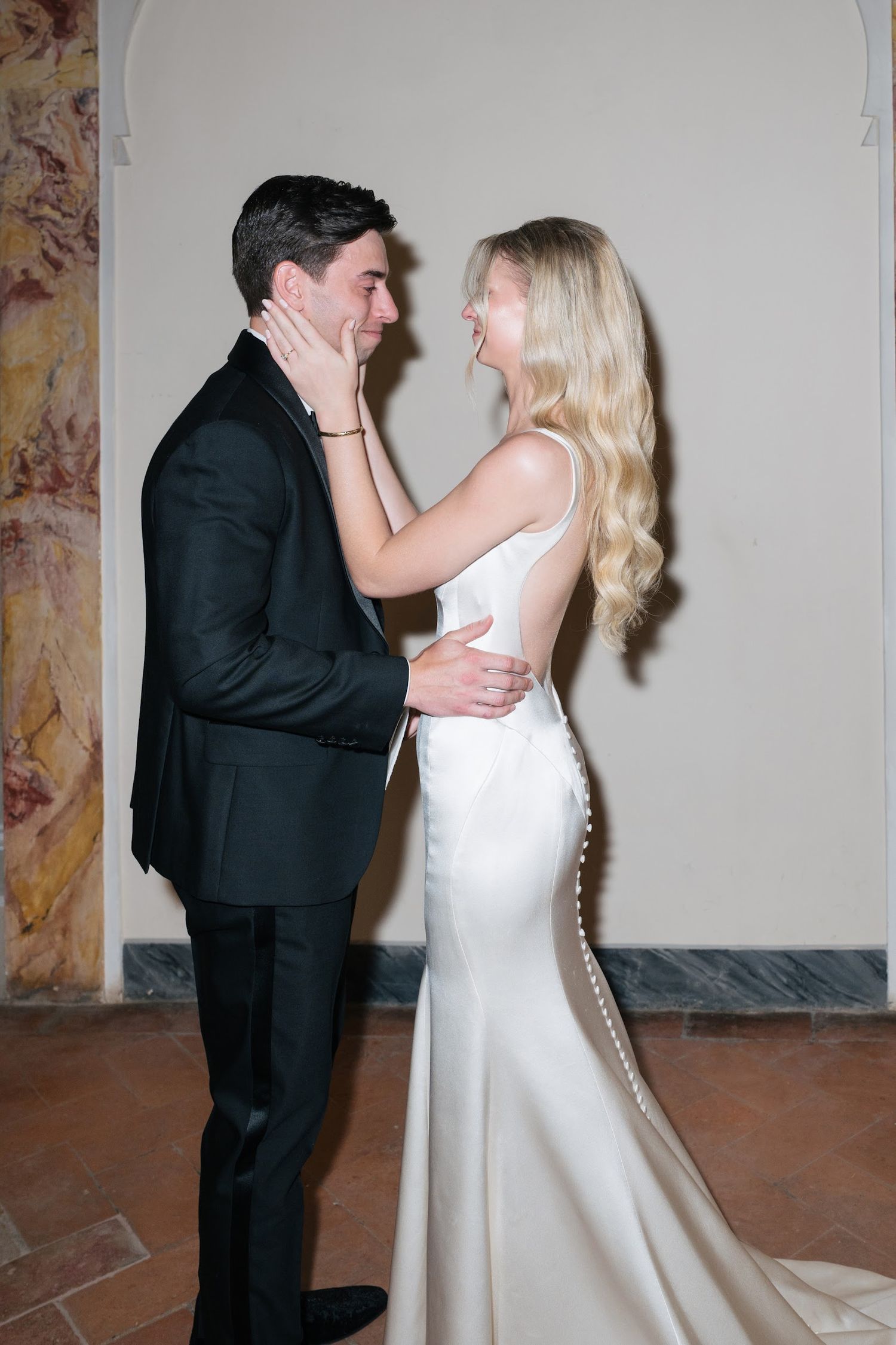A joyful bride in a white satin gown tenderly cups her grooms face during their wedding ceremony.