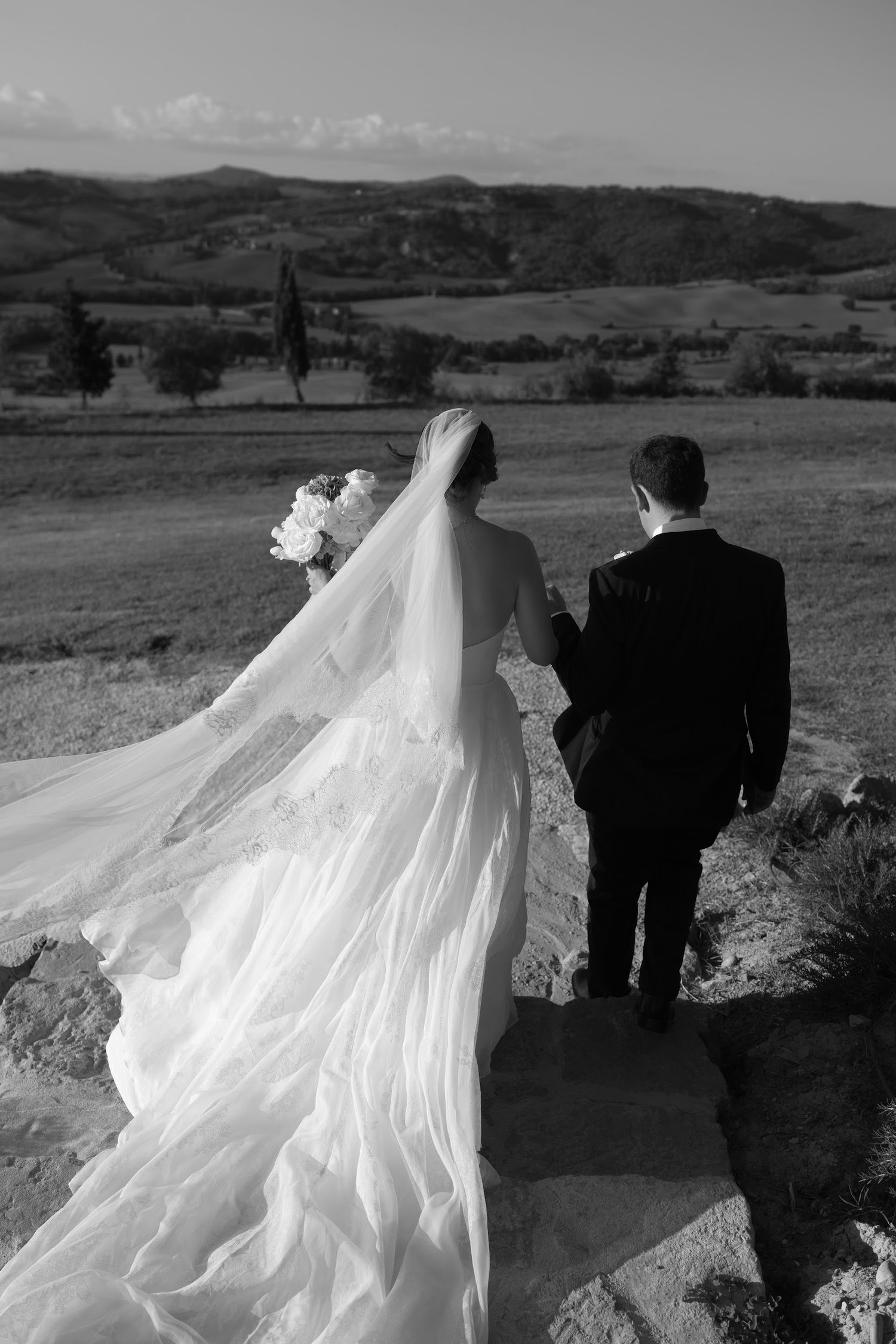 Black and white photo of bride and groom walking with flowing veil in Tuscan landscape.