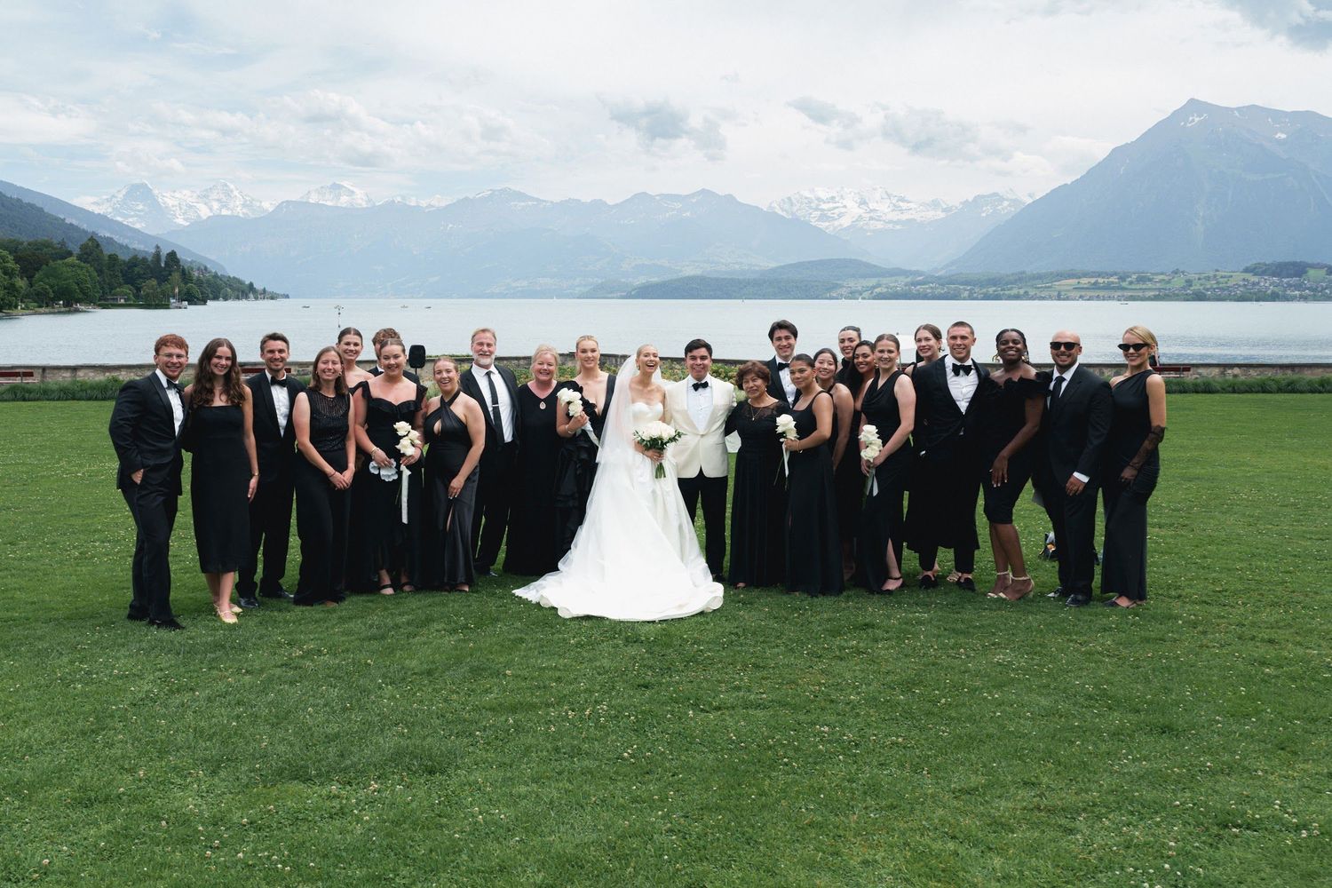 Wedding party group photo on green lawn with bride, groom, and guests in black formal attire against alpine lake scenery.