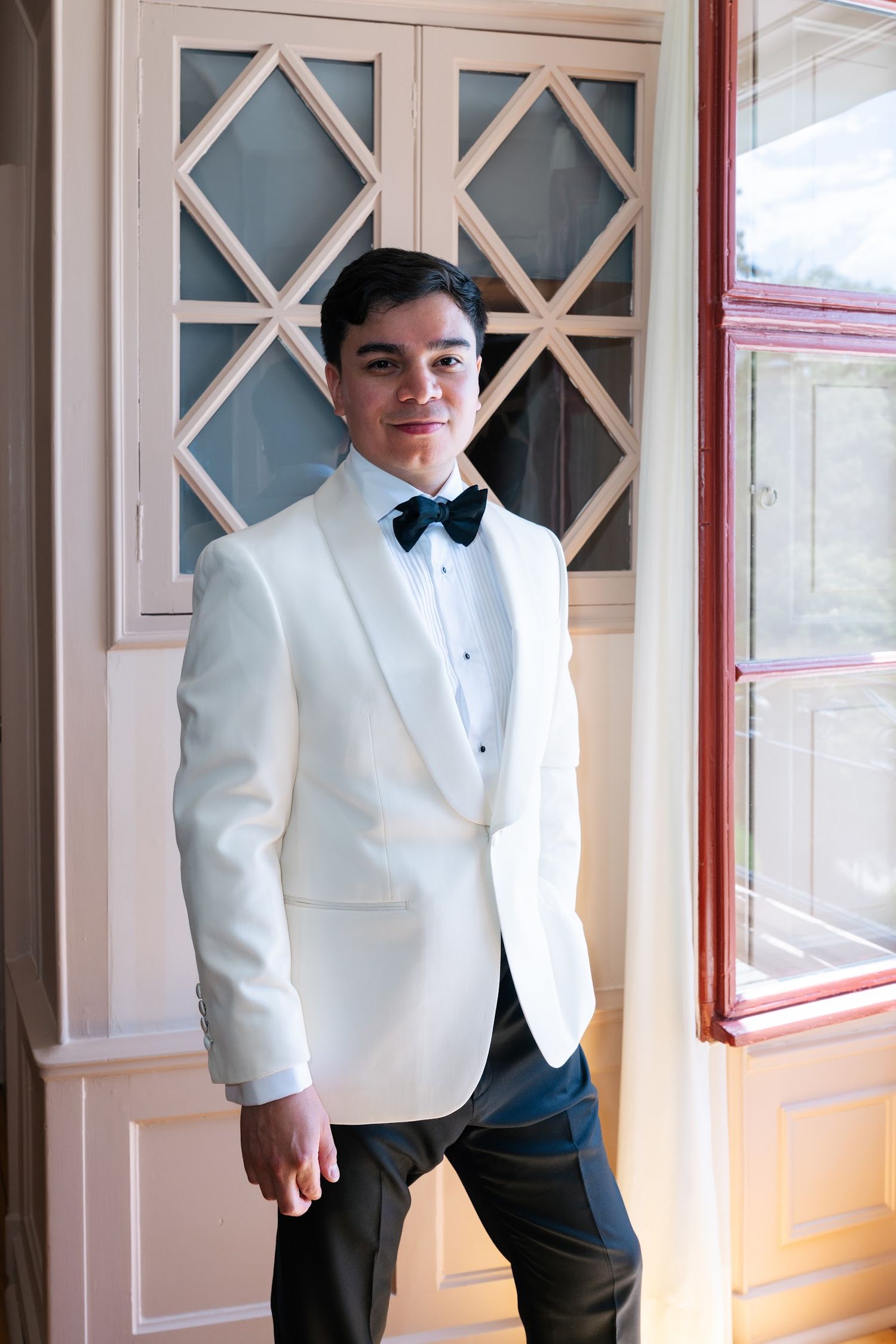 Handsome groom in white tuxedo jacket and black bow tie posing by ornate window doors.