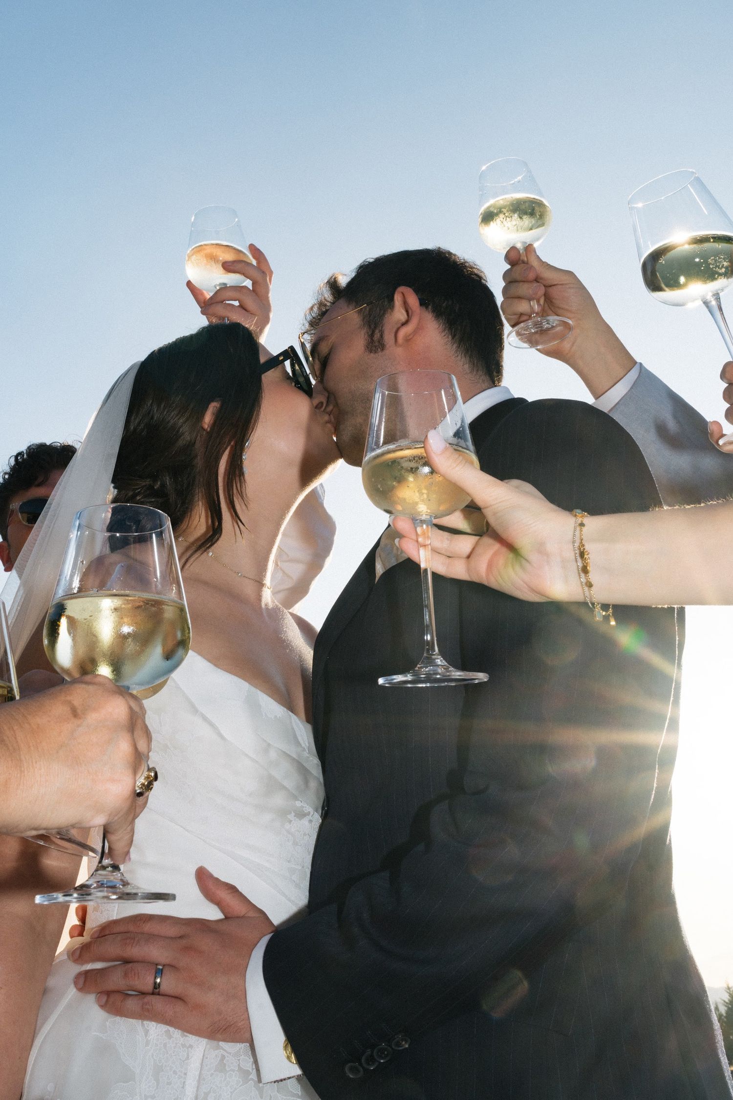 Bride and groom share a kiss while wedding guests raise champagne glasses in a sunny outdoor celebration.