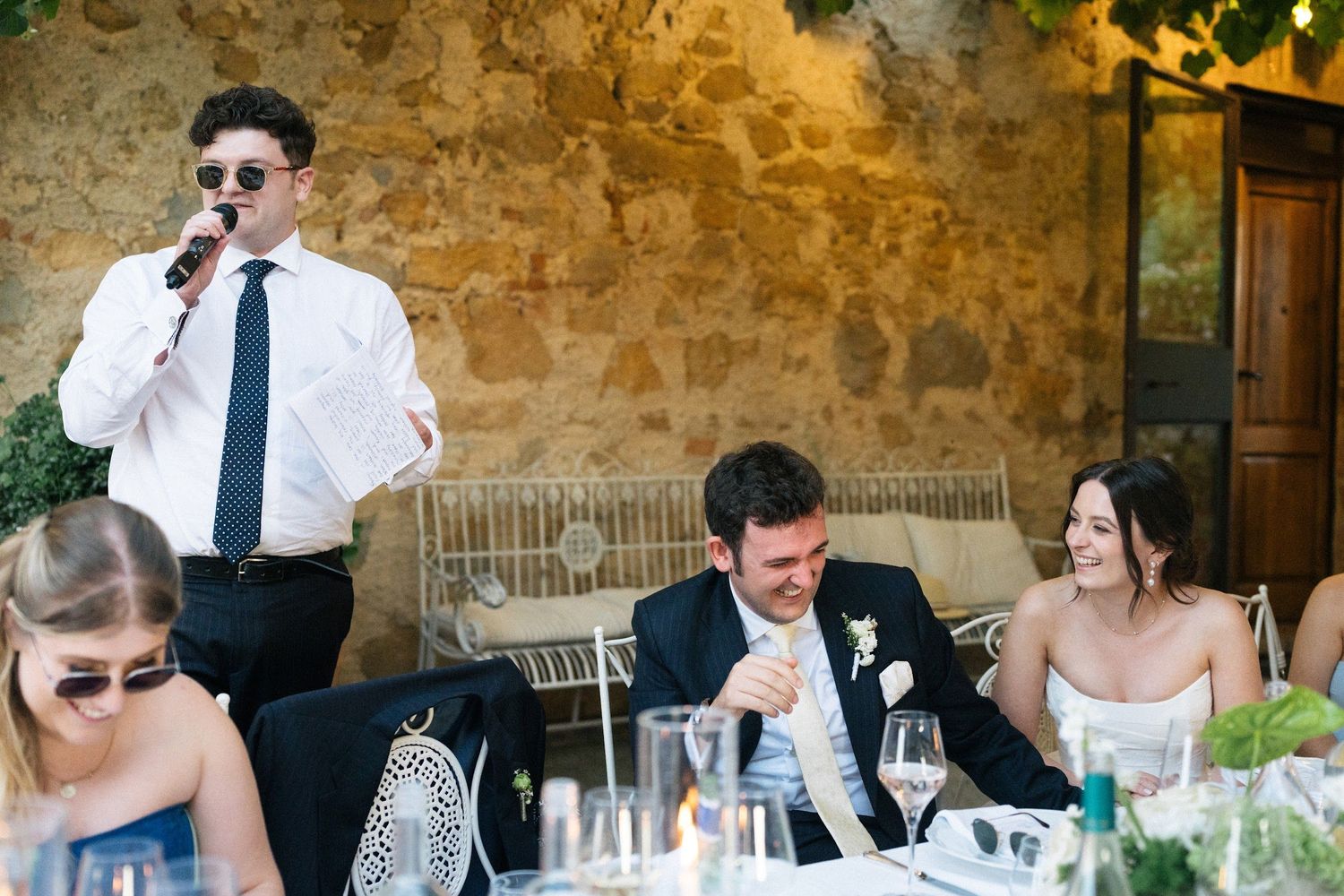 Best man delivers humorous wedding speech into microphone as bride and groom laugh at outdoor reception.