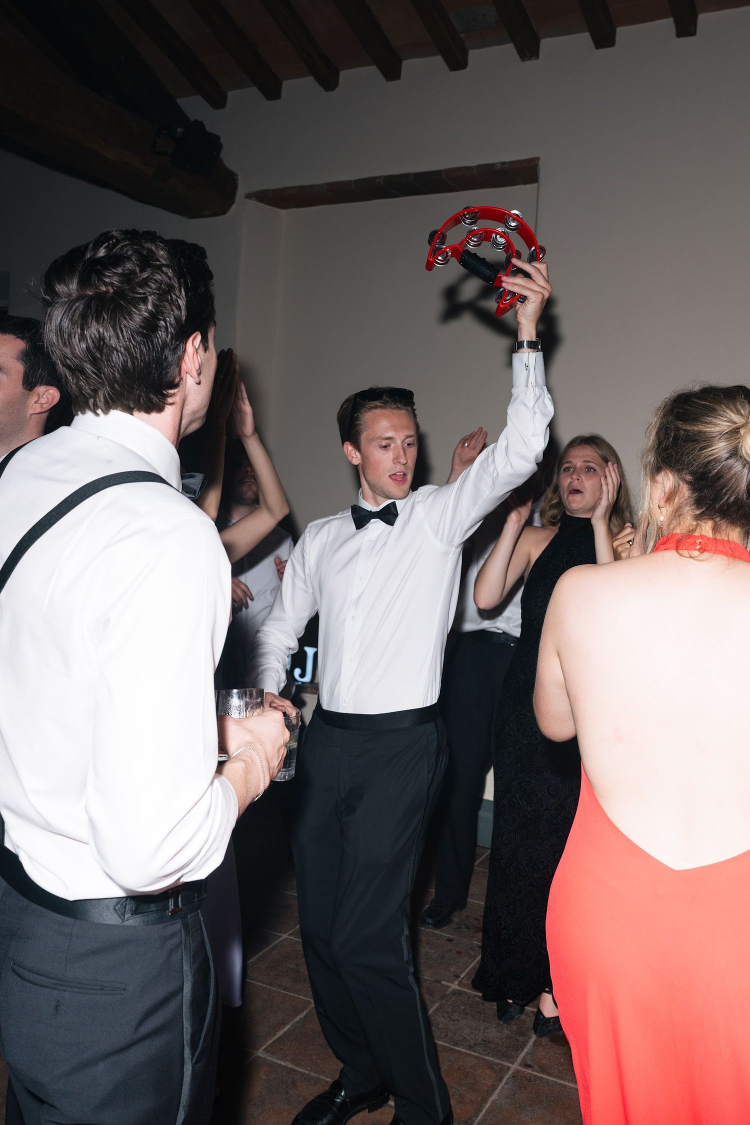 Energetic wedding guest in bow tie raises tambourine while dancing at lively reception.