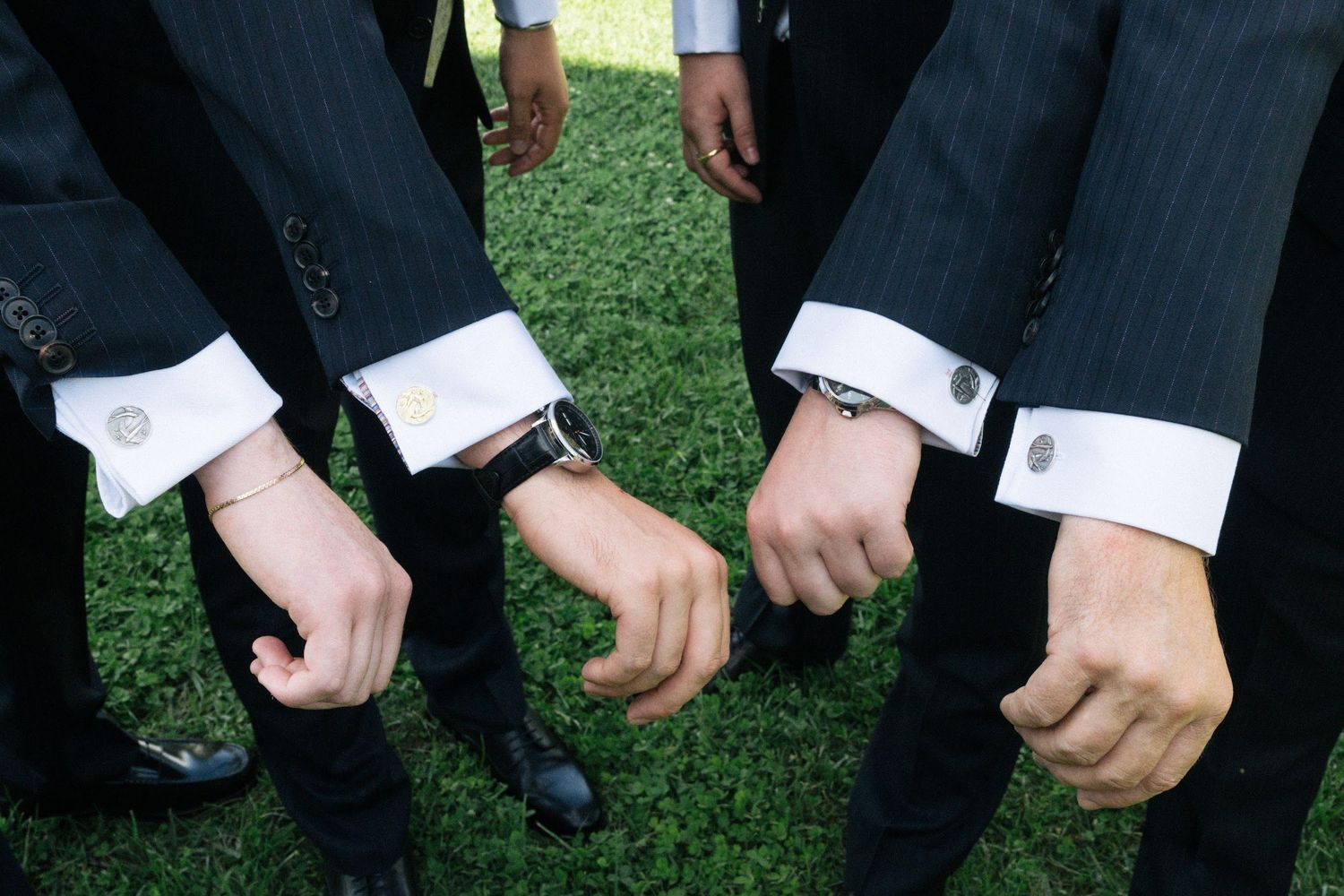 Four groomsmen in dark suits displaying their cufflinks by extending their fists together over green grass.