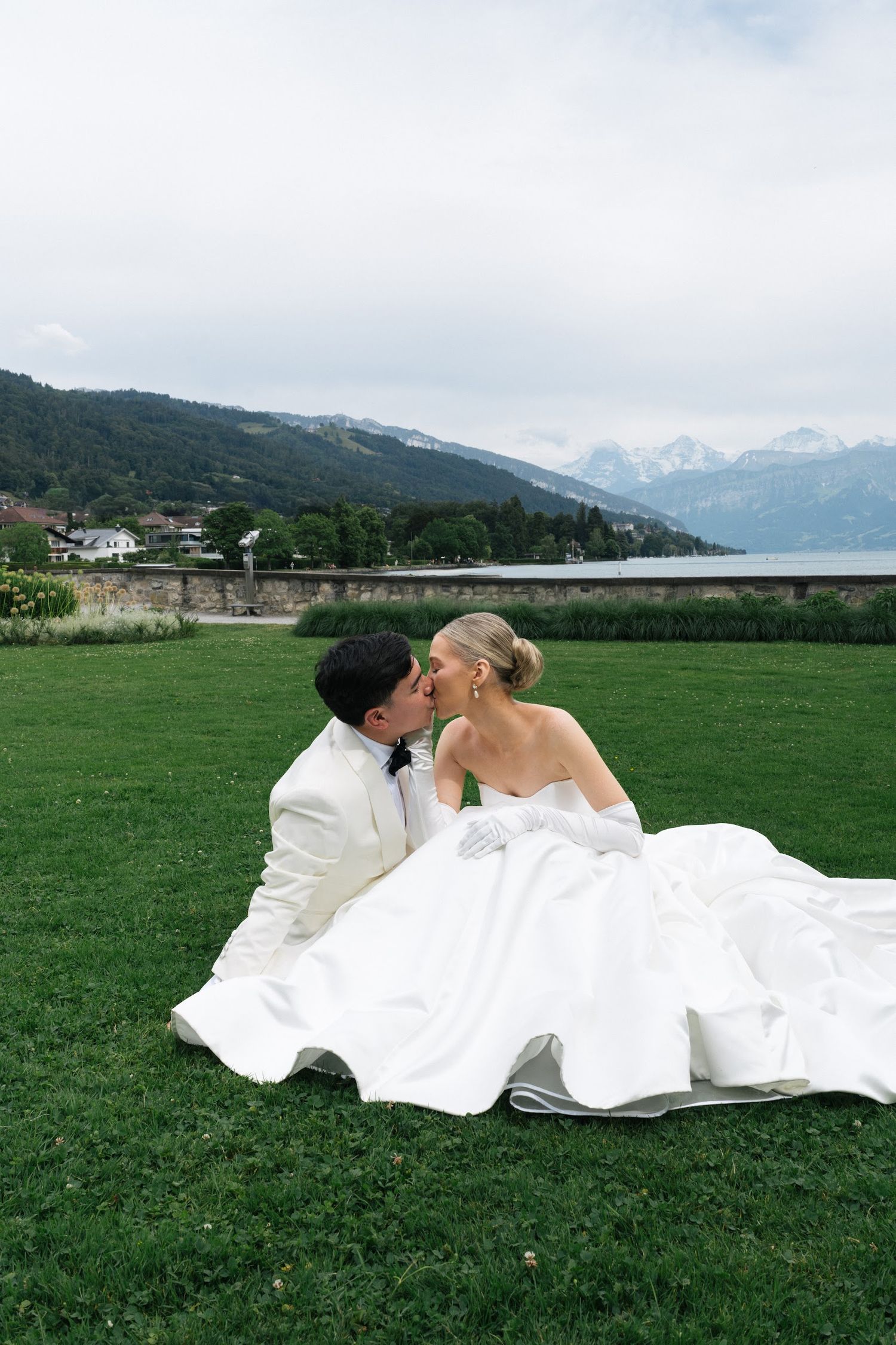 Bride and groom sharing a romantic kiss while seated on grass with alpine lake scenery.
