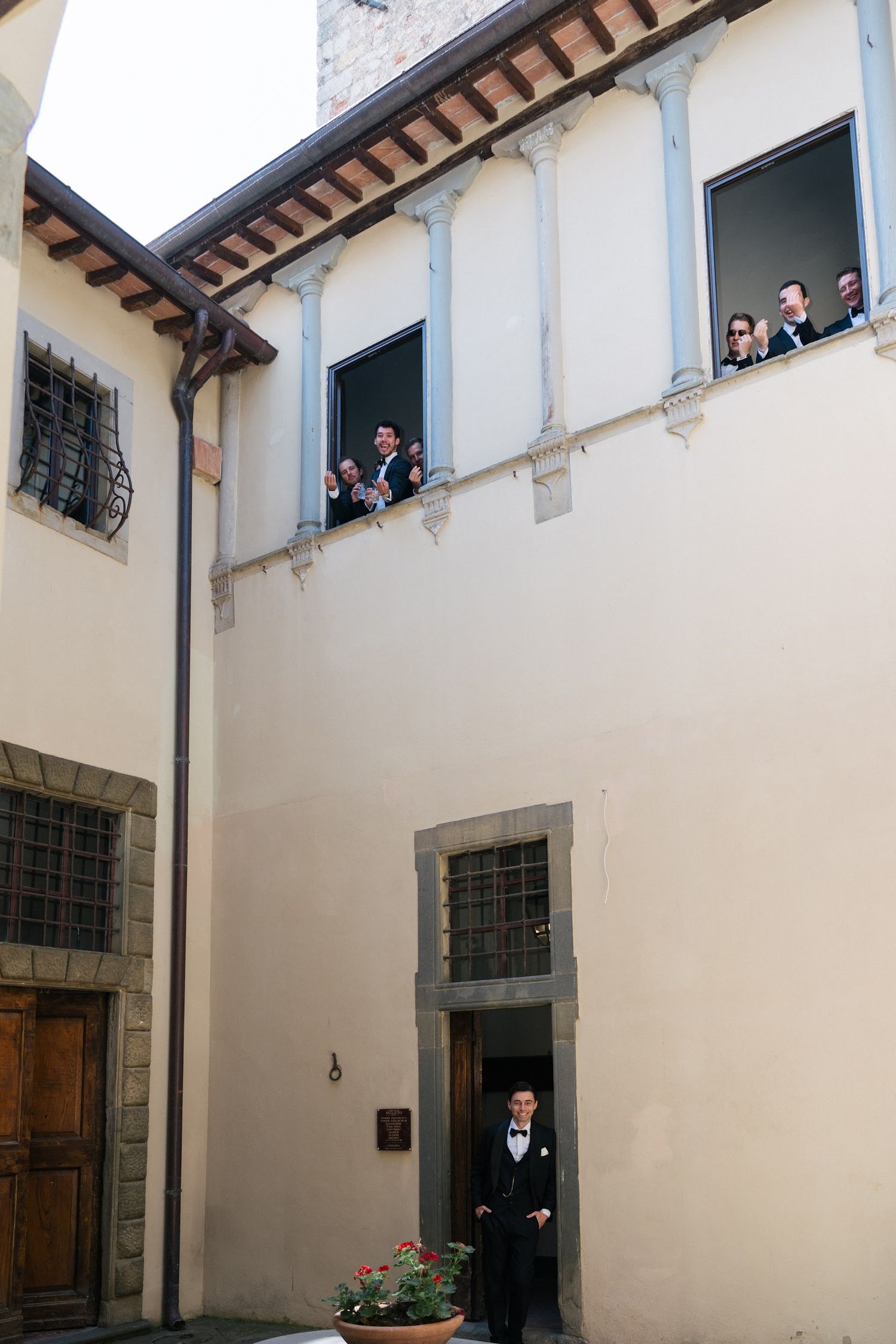 Groom in black tuxedo exits doorway as wedding party watches from upper balcony of Italian villa.
