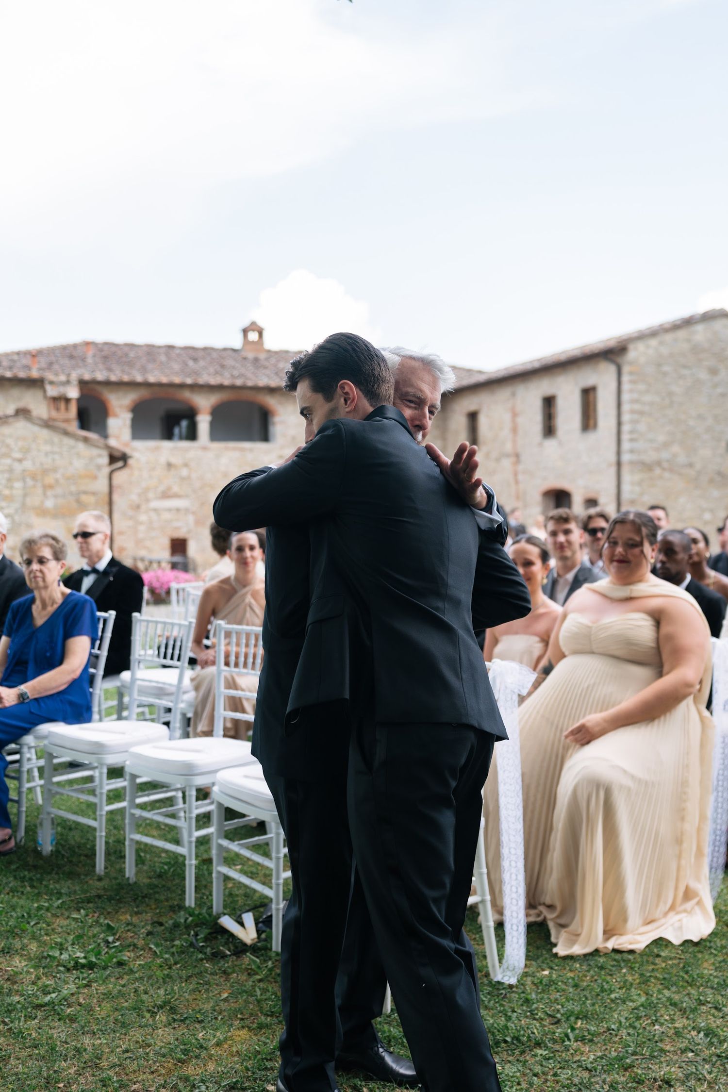 Two men in tuxedos share an emotional embrace during outdoor wedding ceremony at rustic Italian venue.