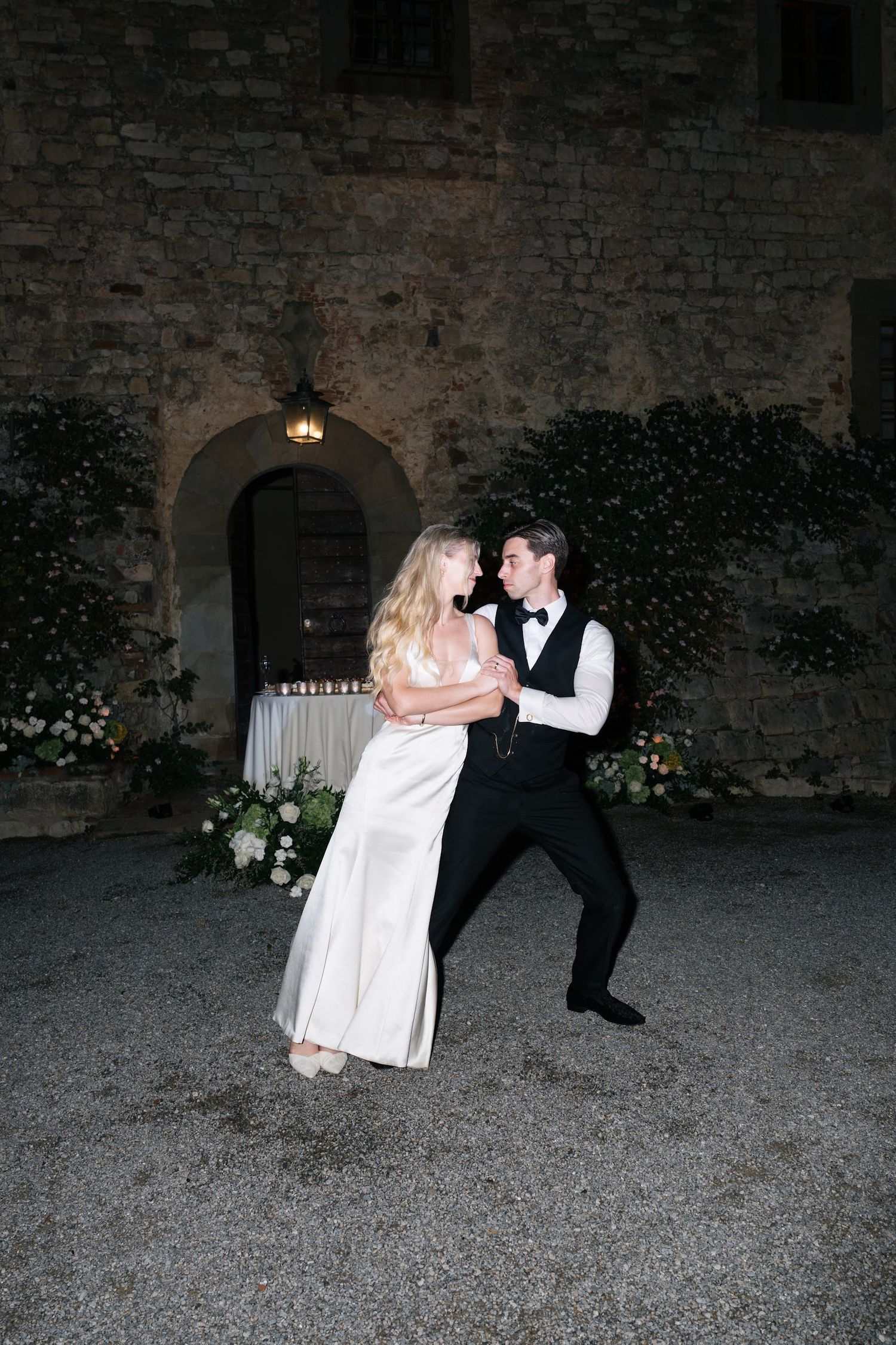 Bride and groom sharing a joyful first dance outside a rustic stone castle venue at night.