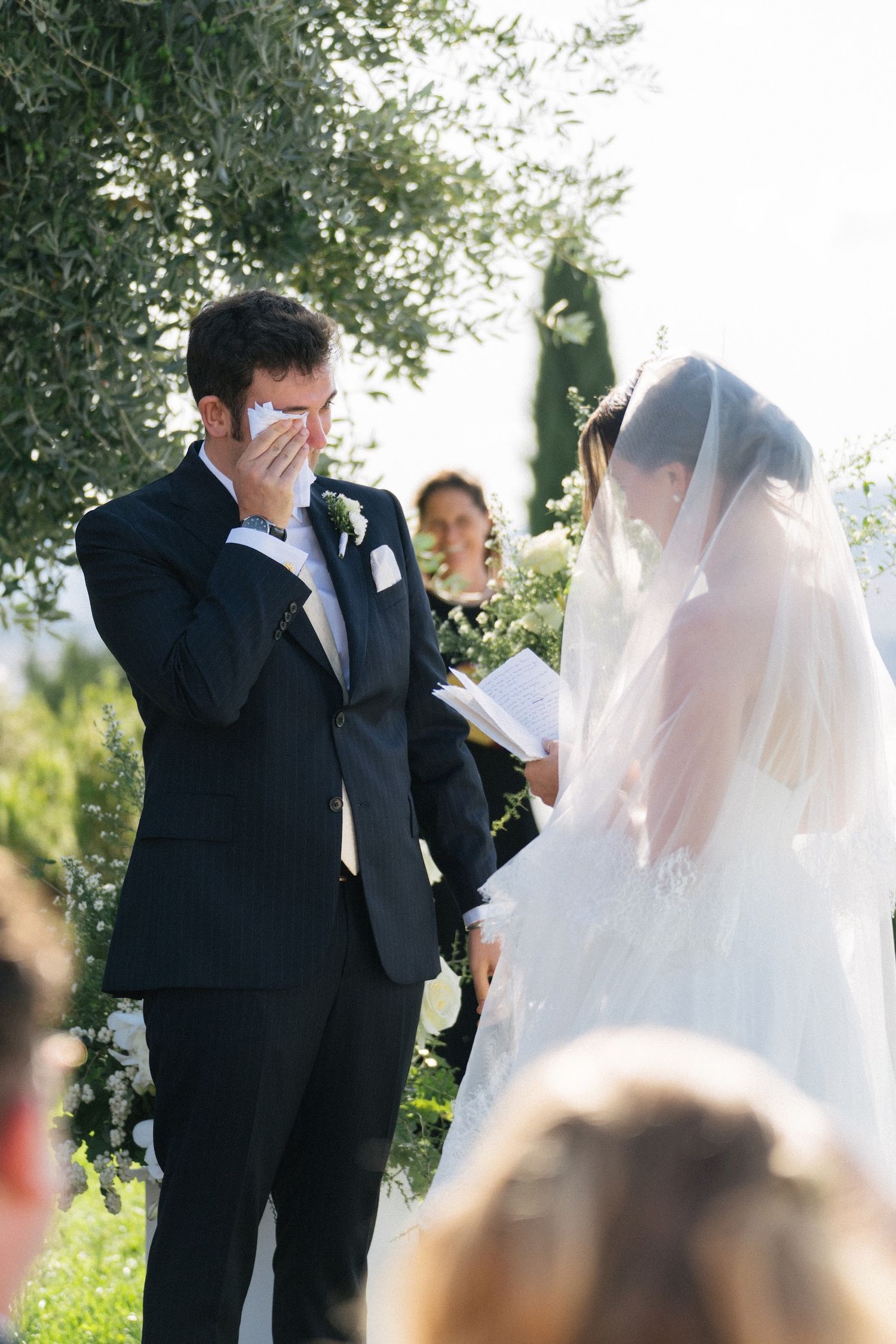 Teary groom wiping eyes as bride reads vows during sunny outdoor wedding ceremony.