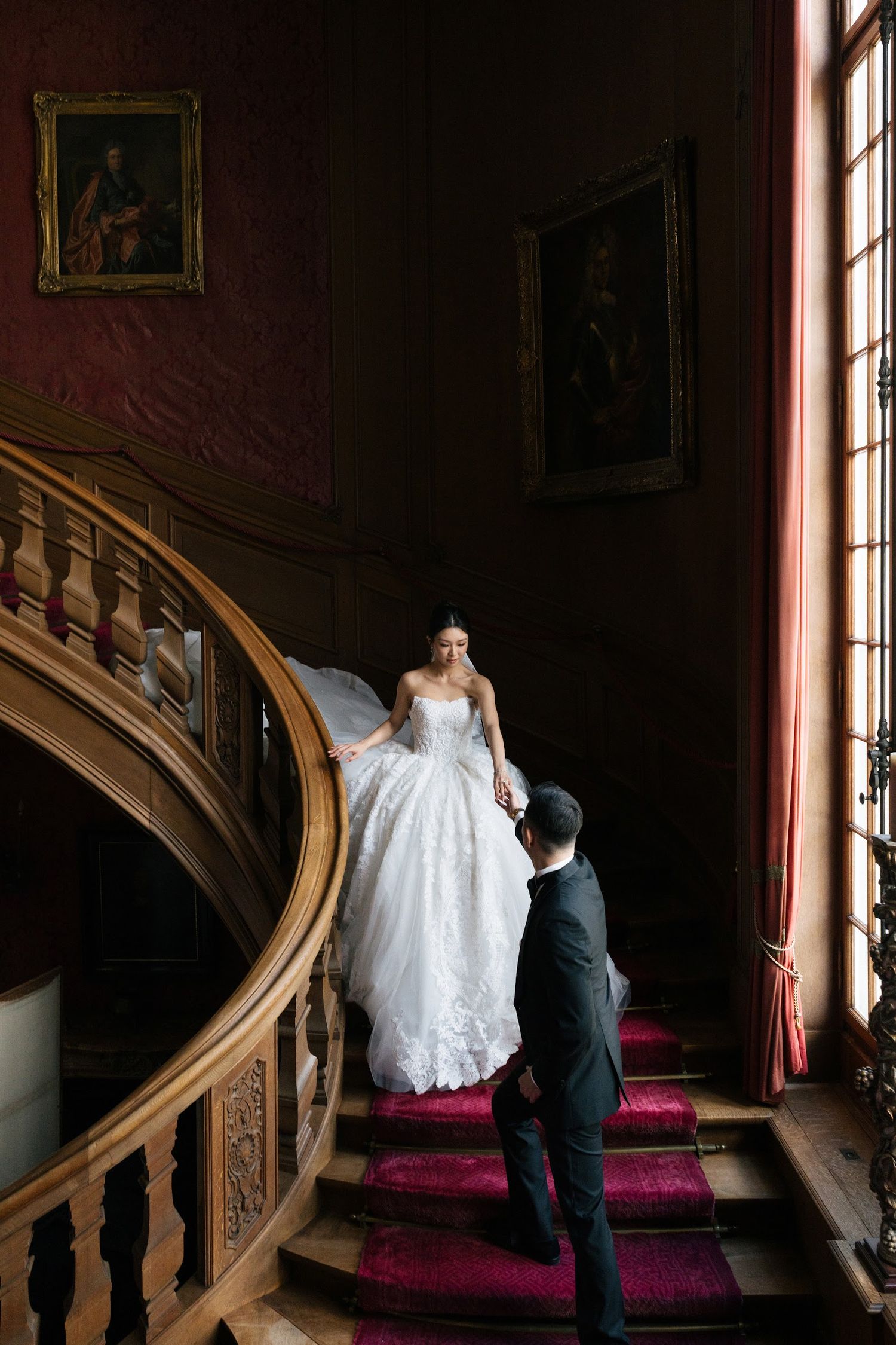 Bride in lace ballgown descending grand wooden staircase with red carpet alongside groom.