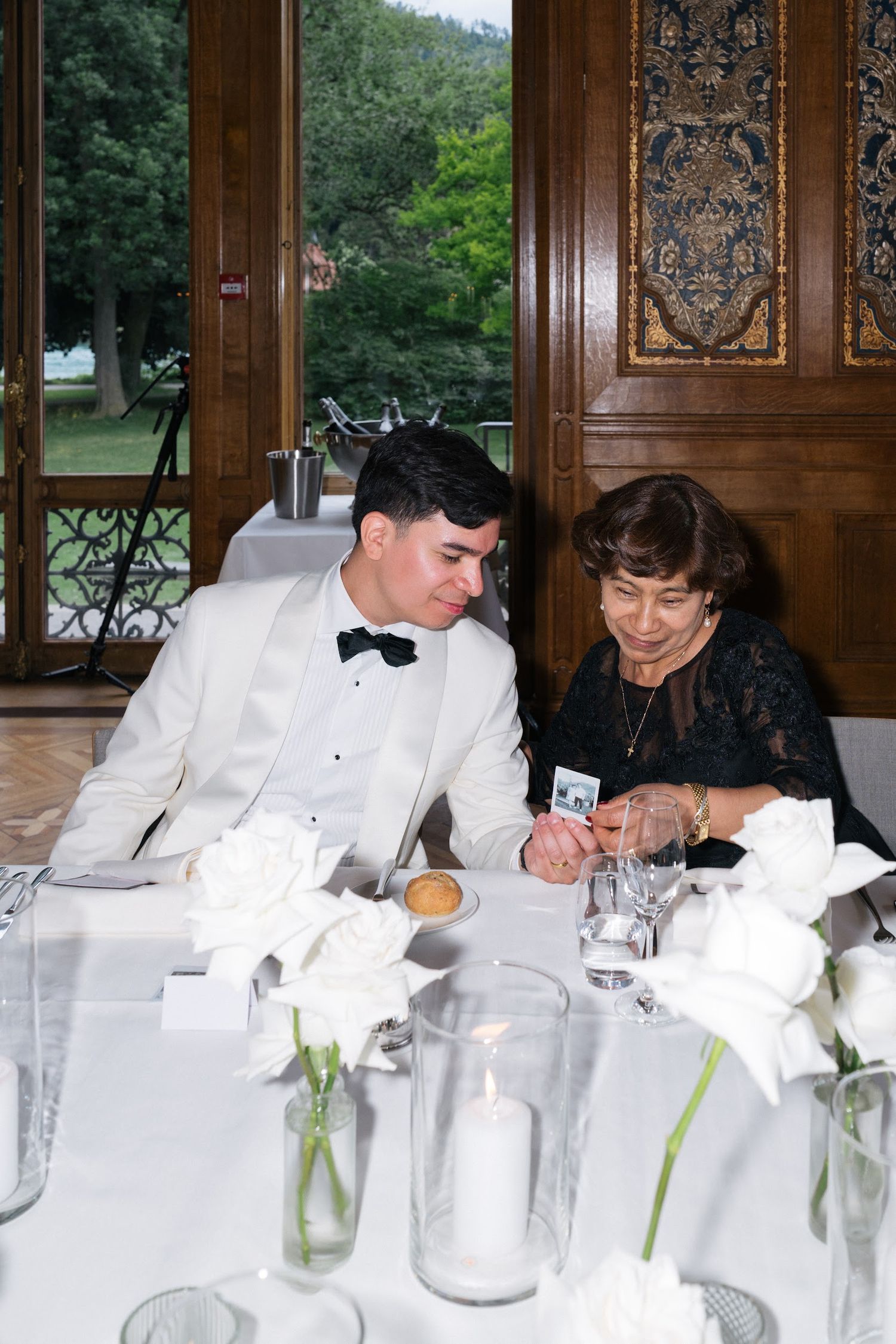 Groom in white tuxedo shares a moment with older female guest at formal wedding dinner table.