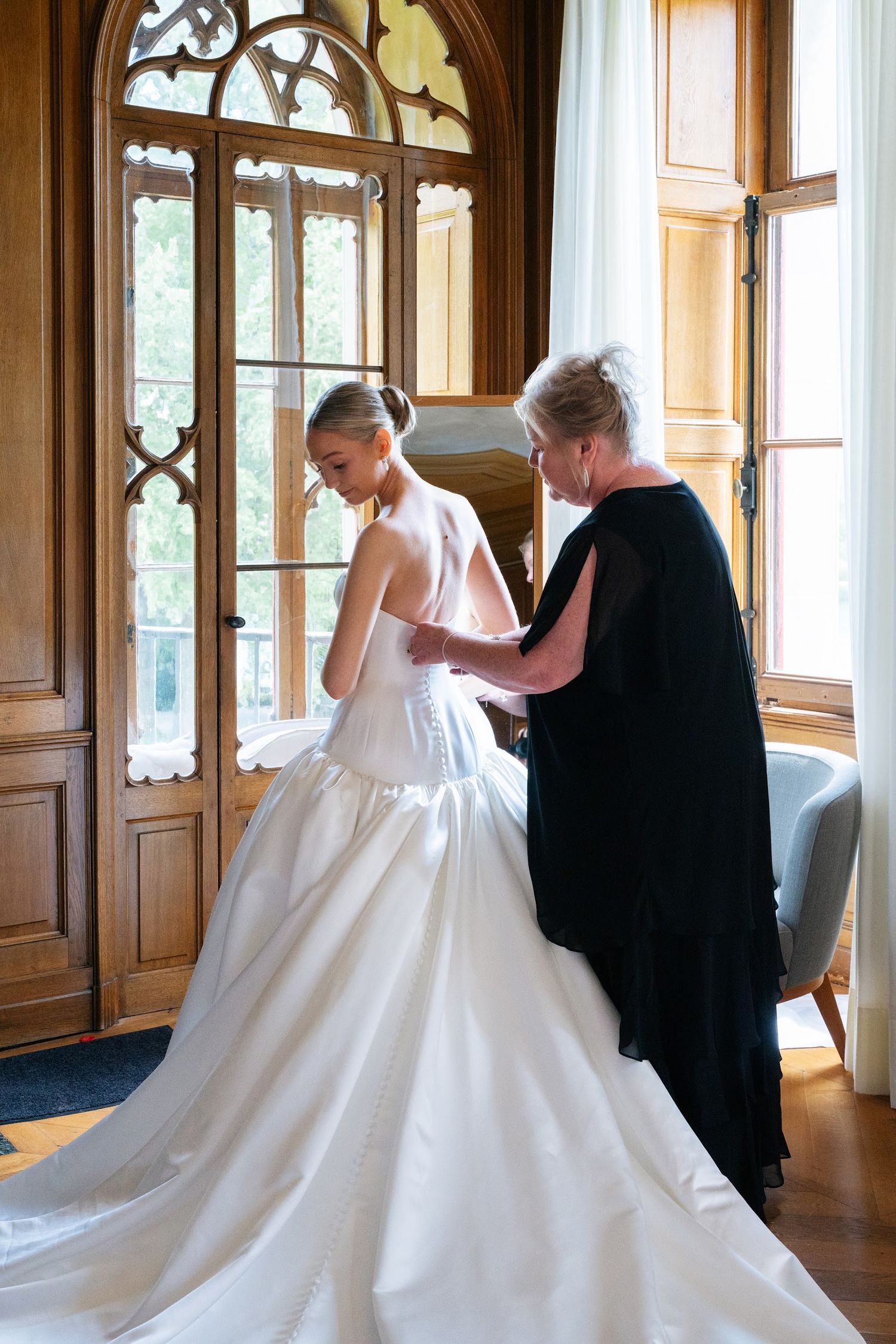 Mother helping bride fasten stunning white ball gown by ornate gothic arched windows.